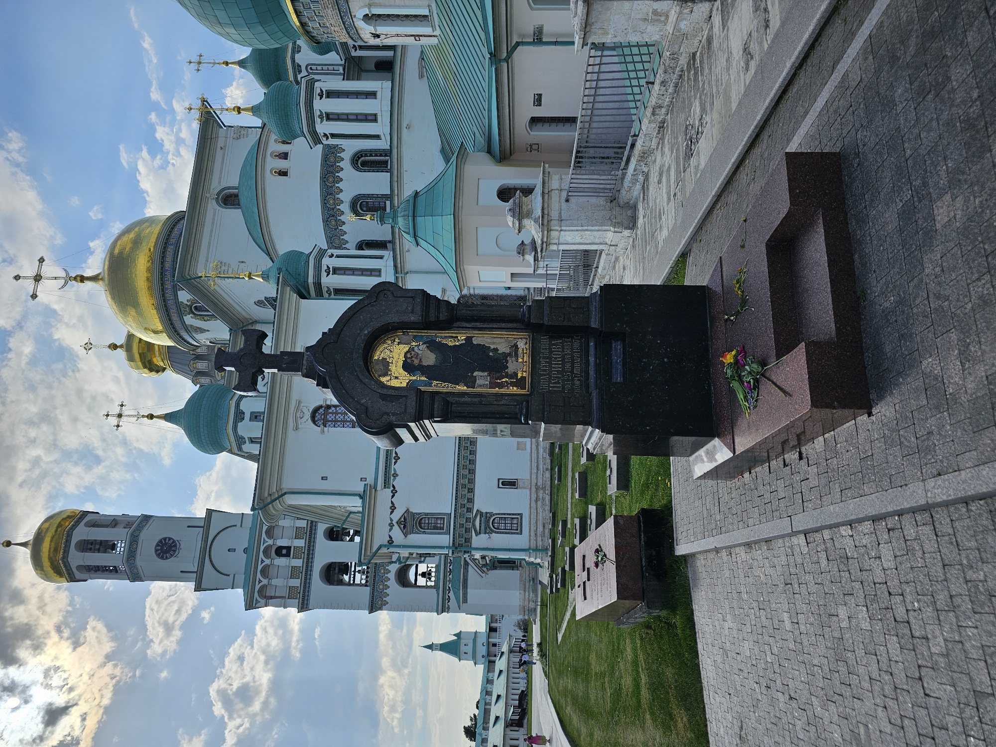 Here is a description of the image:
The image shows a view of the New Jerusalem Monastery in Istra, Russia. The foreground features a black marble gravestone, adorned with a golden religious icon and Cyrillic inscriptions. A red granite monument resembling a cross stands near the gravestone. Flowers have been placed on the graves. In the background, the white walls and turquoise domes of the monastery rise majestically against a partly cloudy sky. The exterior architecture of the monastery showcases intricate details. A paved walkway borders the graves. The composition of the image juxtaposes the personal markers of remembrance with the grandeur of the historical and religious structure.