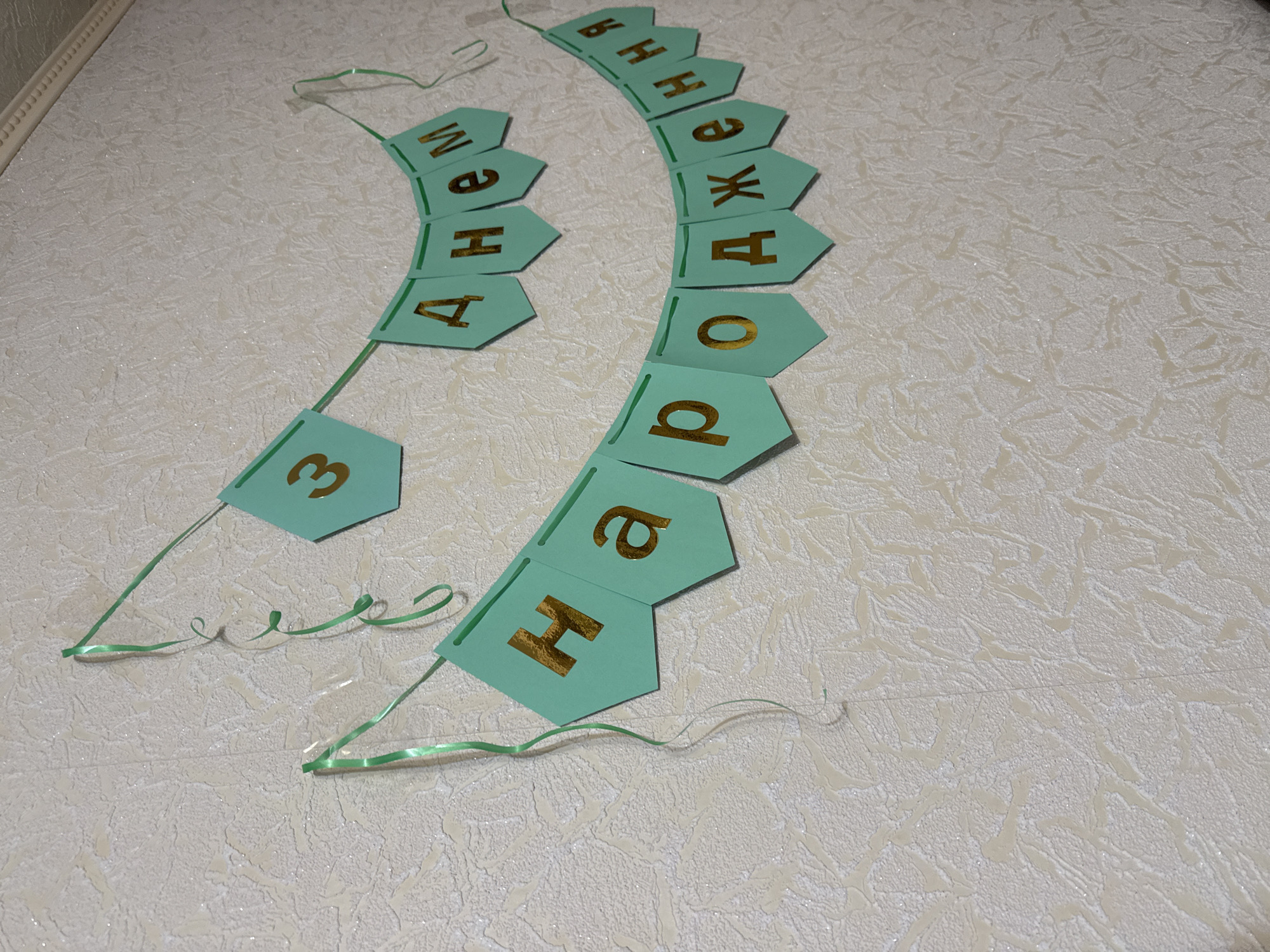 A close-up, eye-level shot shows two strings of pennant banners hanging on a textured, off-white wall. The banners are hexagonal and mint green, with gold foil letters on them. The upper banner reads "З Днем" (Happy Birthday in Ukrainian). The lower banner reads "Народження" (Birthday in Ukrainian). Green ribbons are tied to the top of each banner, with some ribbons curled. The wall has a rough, almost stucco-like texture with some subtle glitter. The lighting is soft, creating some shadows.