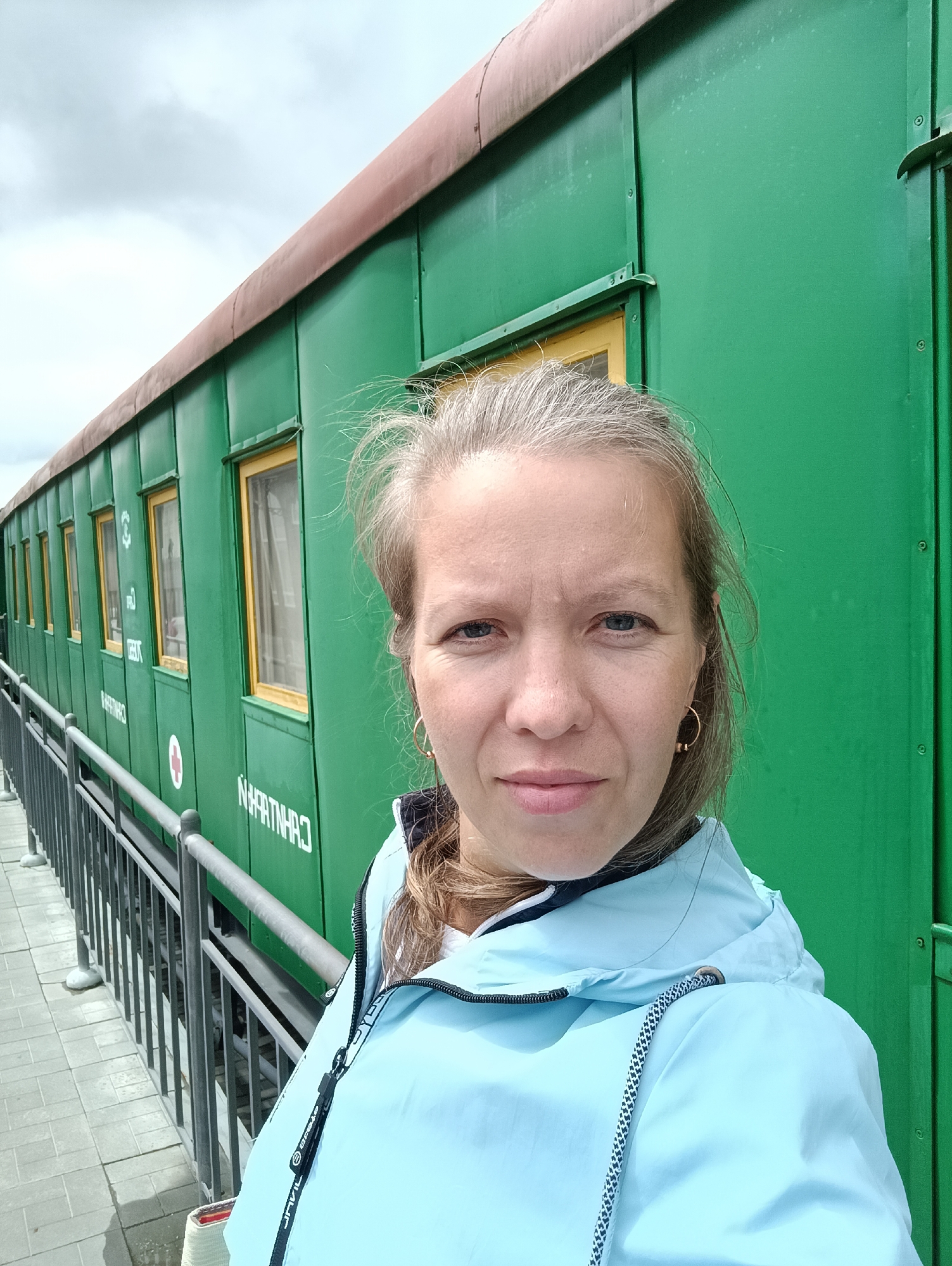 A woman with light brown hair, wearing a light blue hooded jacket, poses for a selfie in front of a green train car. The train car appears to be an old passenger car with several windows and some Russian text visible on its side. A metal railing runs along the side of the train car, separating it from a paved walkway. The sky in the background is overcast. The train car is likely on display at the Military Technology Museum in Verkhnyaya Pyshma, Russia, given the museum's collection of historical military equipment, which includes trains.