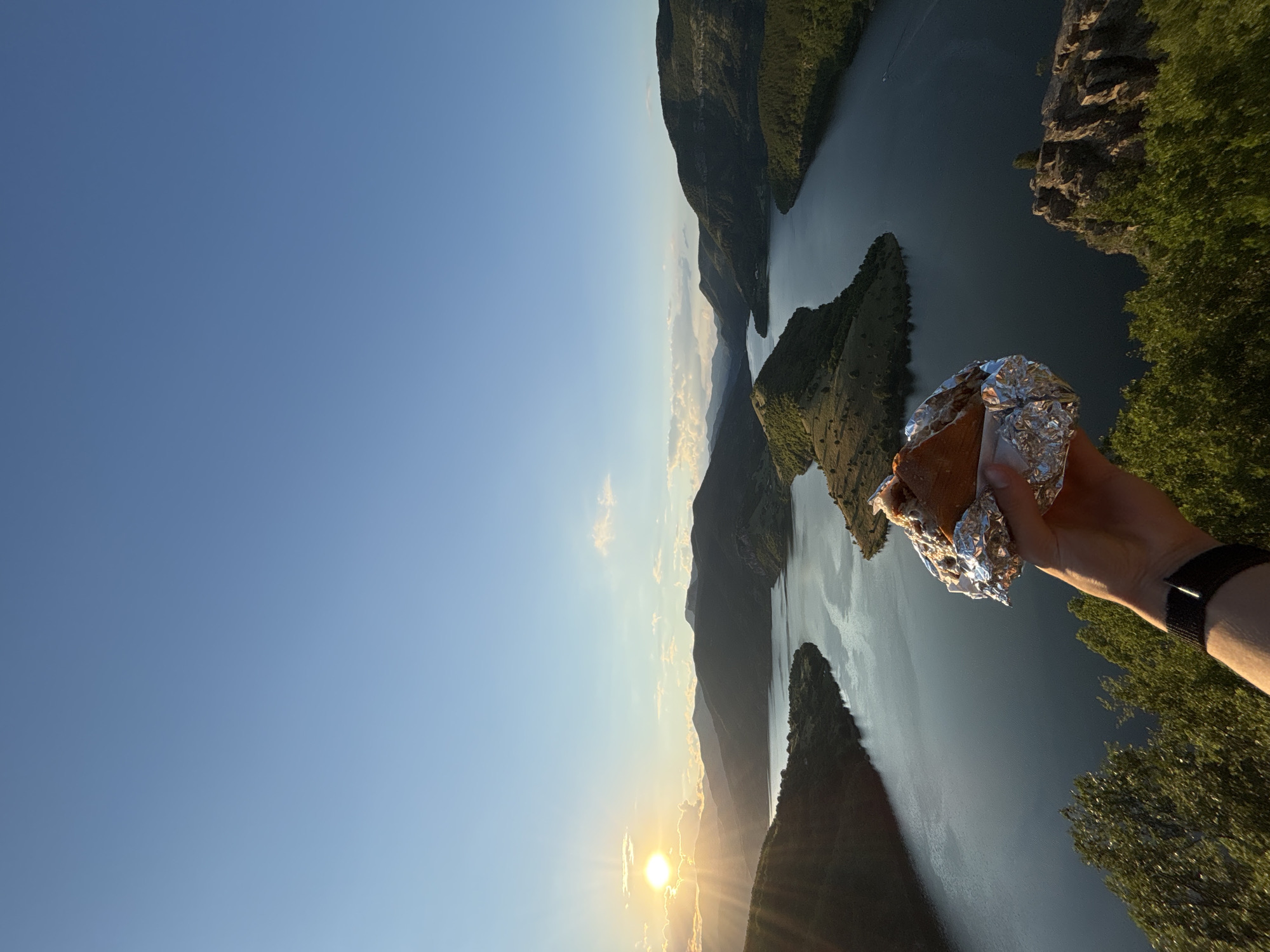 In the foreground, a hand holding a sandwich wrapped in aluminum foil is positioned in front of a scenic vista. The sandwich appears to be partially unwrapped, revealing a pastry with a filling. The hand wears a black watch on the wrist. Behind the hand and sandwich, a wide, dark blue lake winds through a valley surrounded by lush green, rolling hills and rugged mountains. The sun is setting on the left side of the image, casting a warm, golden glow and long shadows across the landscape. Rays of sunlight break through scattered clouds, illuminating parts of the hills and the water. The sky is a clear, pale blue, with a few wispy clouds. In the distance, more mountains fade into the horizon. A small boat is visible on the lake. The scene is a panoramic view of a natural landscape, likely in the vicinity of Borovitsa, Bulgaria, given the distinctive horseshoe-shaped bend of the Arda River, often referred to as the "Borovitsa Horseshoe."