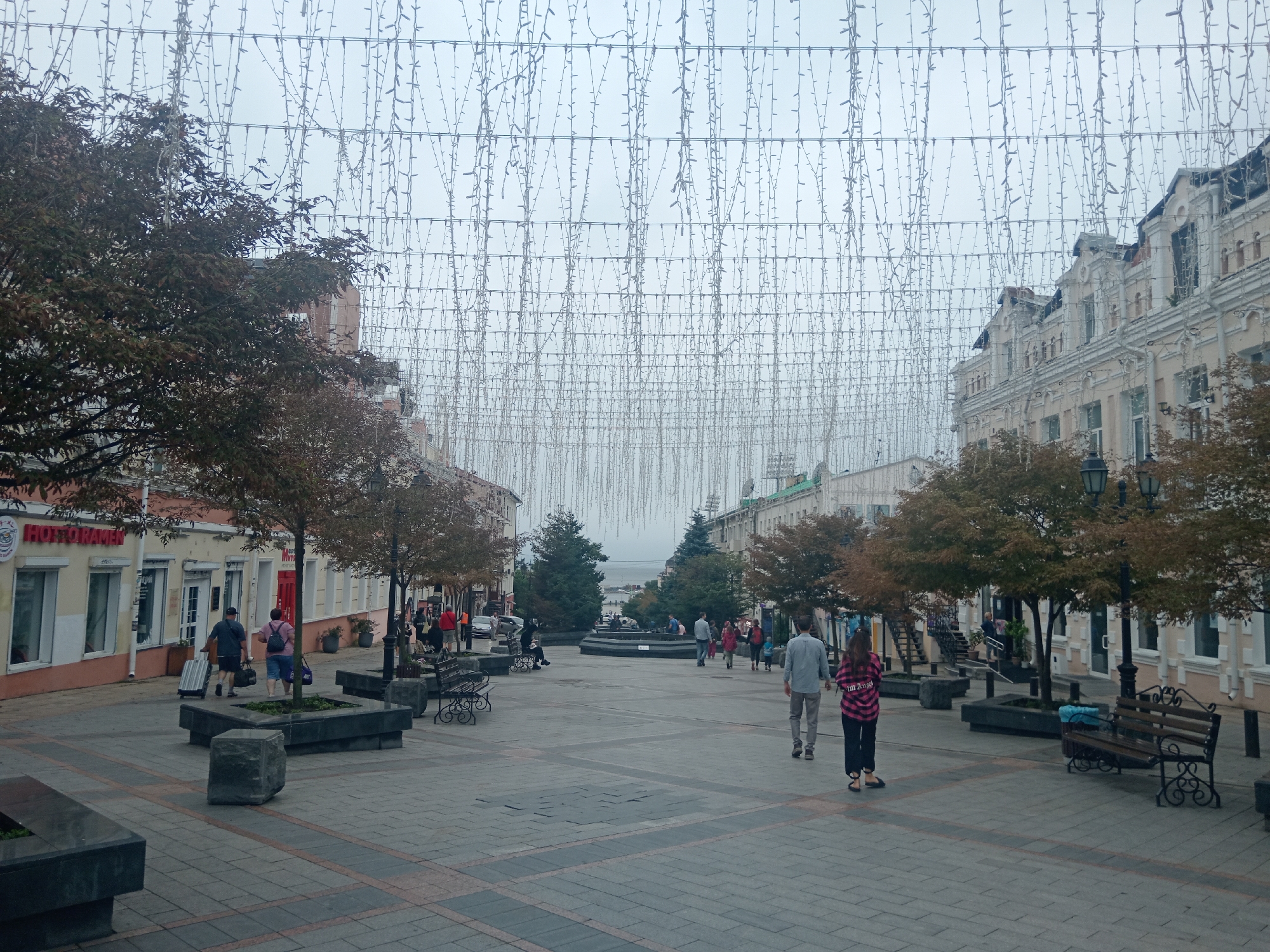 This image depicts a pedestrian street in Vladivostok, Russia, possibly Svetlanskaya Street, characterized by an overcast sky and overhead string lights. The street is paved with gray and red-toned cobblestones, with several planters made of dark stone. On the left, buildings with light-colored facades and prominent signage are visible, including one with "HOT RAMEN" in English. A few trees with sparse, autumnal foliage are planted along the sidewalk.
Several individuals are present. Two people walk away from the camera on the left, one pulling a suitcase. Further down the street, groups of people are scattered, some walking, some sitting on benches. On the right, a light-colored, ornate building with multiple stories and arched windows lines the street. A black wrought-iron bench is visible in the foreground on the right. The scene appears to be a public square or a pedestrian zone within the city.