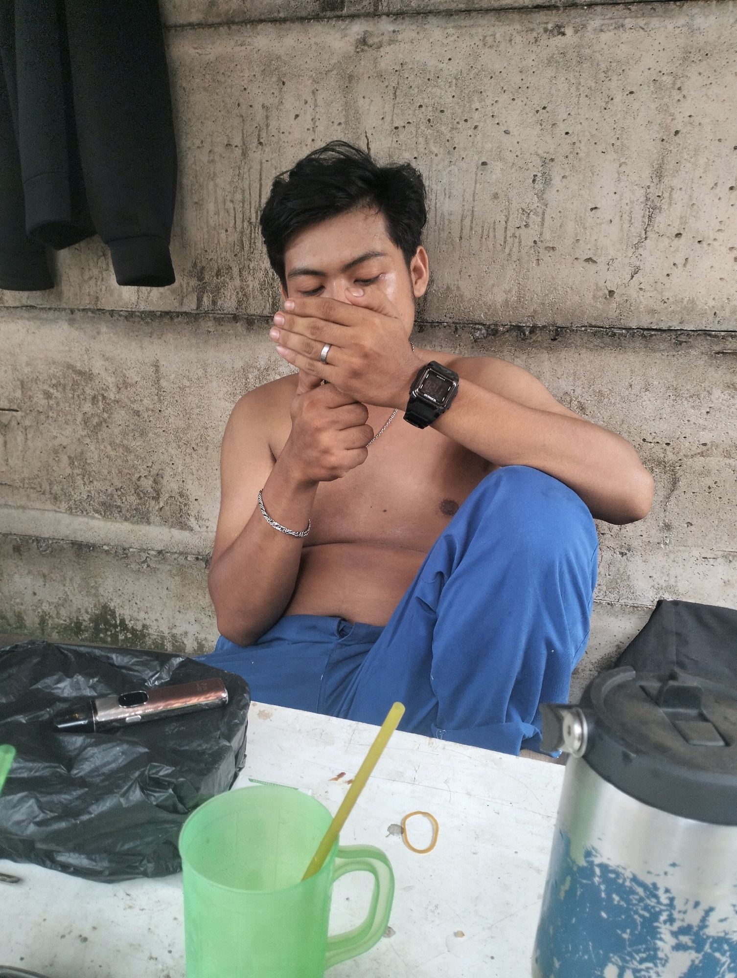 A young shirtless man with dark hair, wearing blue pants and a black watch on his left wrist, sits on a concrete ledge against a weathered concrete wall. His right hand is covering his mouth, and a silver ring is visible on his left ring finger. To his left, on a white surface, is a black plastic bag containing a metallic vape pen. A green plastic cup with a yellow straw is also on the white surface. To the right of the frame, a metallic thermos with blue paint splatters is partially visible. A black garment hangs on the wall behind the man. The scene appears to be outdoors, possibly in a public area in Pontianak, Indonesia.