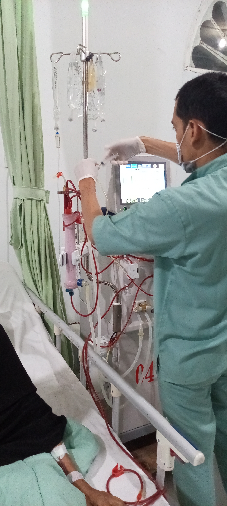 The image shows a medical setting, potentially in a hospital or dialysis center. A healthcare worker, wearing a light green scrub suit, a medical mask, and latex gloves, is interacting with a hemodialysis machine. The machine is connected to a patient lying in a bed to the left of the frame. The patient's left arm is visible with a catheter inserted. An IV stand with multiple fluid bags is positioned near the dialysis machine. A curtain drapes in the background. The machine has the text "C4" on it. The location is most likely Lhokseumawe, Indonesia. The healthcare worker appears to be drawing a sample from the dialysis circuit using a syringe.
