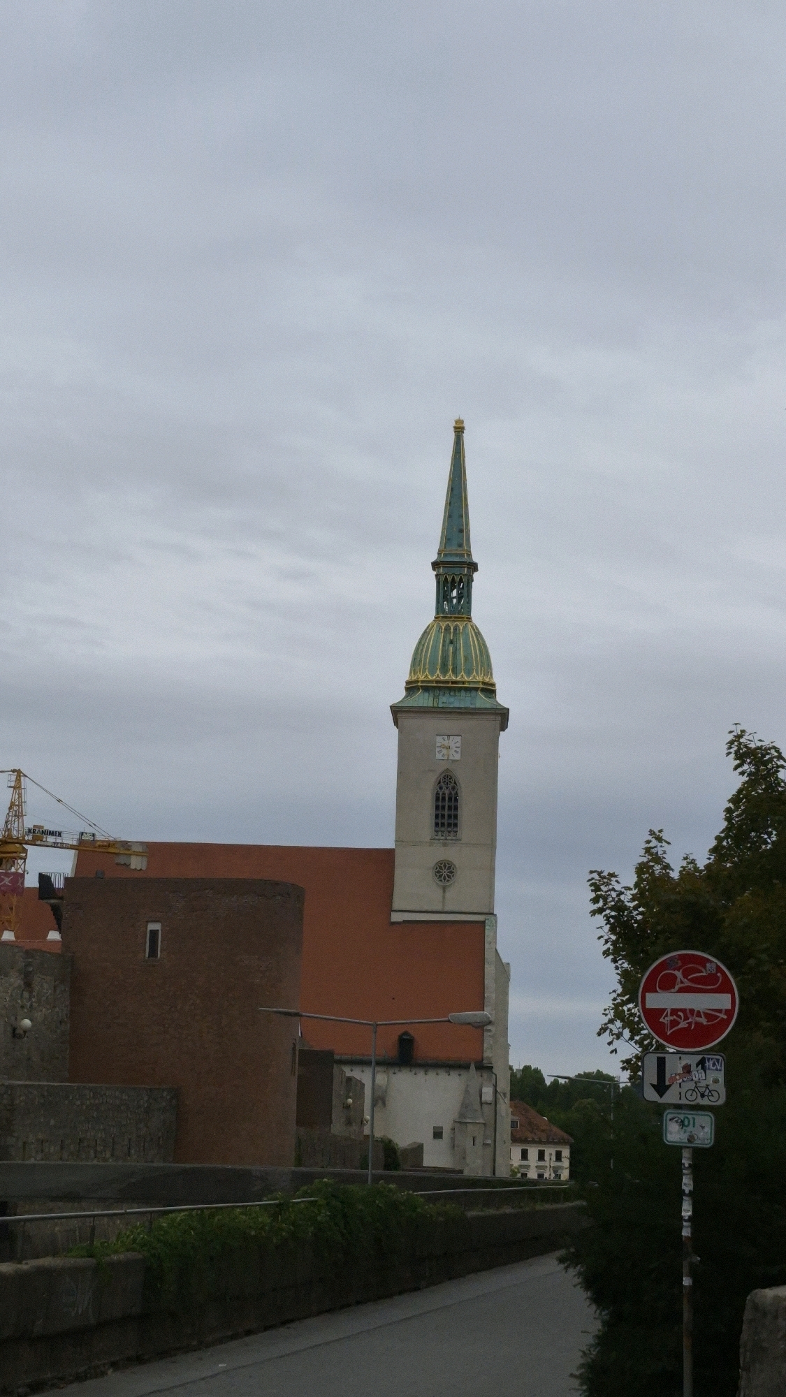 This image captures an exterior view of St. Martin's Cathedral in Bratislava, Slovakia, under an overcast sky. The cathedral's distinctive tall, green and gold spire dominates the upper portion of the frame. The main body of the cathedral features a terracotta-tiled roof and a light-colored tower with a clock face and Gothic-style windows. To the left of the cathedral, a cylindrical, brick-red structure with a small rectangular window is visible, possibly part of the old city walls. In the foreground, a road curves to the right, bordered by concrete barriers and overgrown vegetation. A traffic signpost with multiple signs, including a "no cycling" symbol and directional arrows, stands near the right edge of the frame. Construction cranes are partially visible in the background to the left, suggesting ongoing development or renovation in the area. The overall scene is one of urban architecture set against a muted, cloudy backdrop.