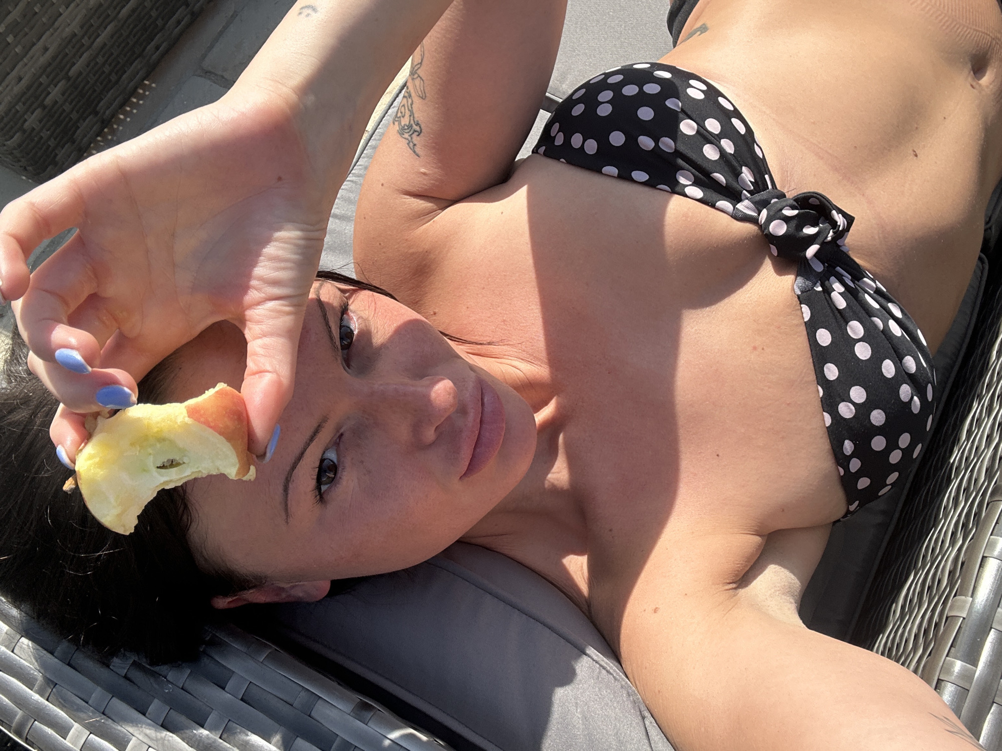 A light-skinned woman lies on a gray cushioned wicker lounge chair, wearing a black and white polka dot bikini top. She holds a partially eaten apple slice near her face with her left hand, which has light blue nail polish. The background shows a brick wall and outdoor furniture, suggesting a patio or garden. The image is brightly lit with sunlight. The location is likely somewhere in Tunbridge Wells, United Kingdom, given the prompt's instructions.