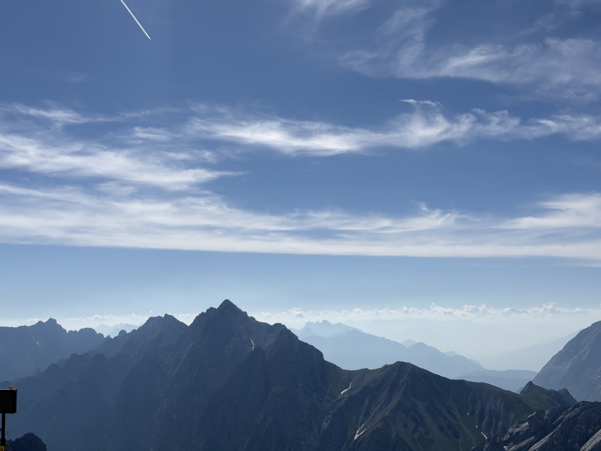 The image depicts a panoramic view of mountain ranges under a bright blue sky with scattered white clouds and a contrail from an airplane. In the foreground, a dark, jagged mountain peak dominates the lower left of the frame. To the left of this peak, a small, dark signpost with a yellow warning symbol is visible. The middle ground features several layers of receding mountain peaks, fading into a hazy blue, suggesting a vast distance. The background is an expanse of sky, with a prominent horizontal band of thinner, wispy clouds stretching across the mid-horizon. The lighting suggests it is daytime, likely midday, with the sun casting shadows on the mountains. The overall scene is a natural landscape, likely from a high vantage point. Based on the provided context about Garmisch-Partenkirchen, Germany, this image could represent a view from the Zugspitze, Germany's highest peak, looking out over the surrounding Alps.