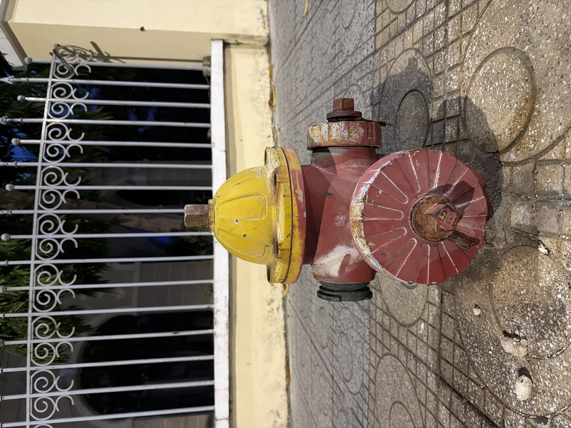 In Nha Trang, Vietnam, a red and yellow fire hydrant stands on a paved sidewalk. The sidewalk is made of concrete tiles with a circular pattern. Behind the hydrant, a decorative metal fence runs along the property line, with a yellow wall and a building visible in the background. The scene appears to be outdoors, possibly in a residential or commercial area, at dusk or dawn, as evidenced by the lighting and shadows. There are no people or animals present in the image.