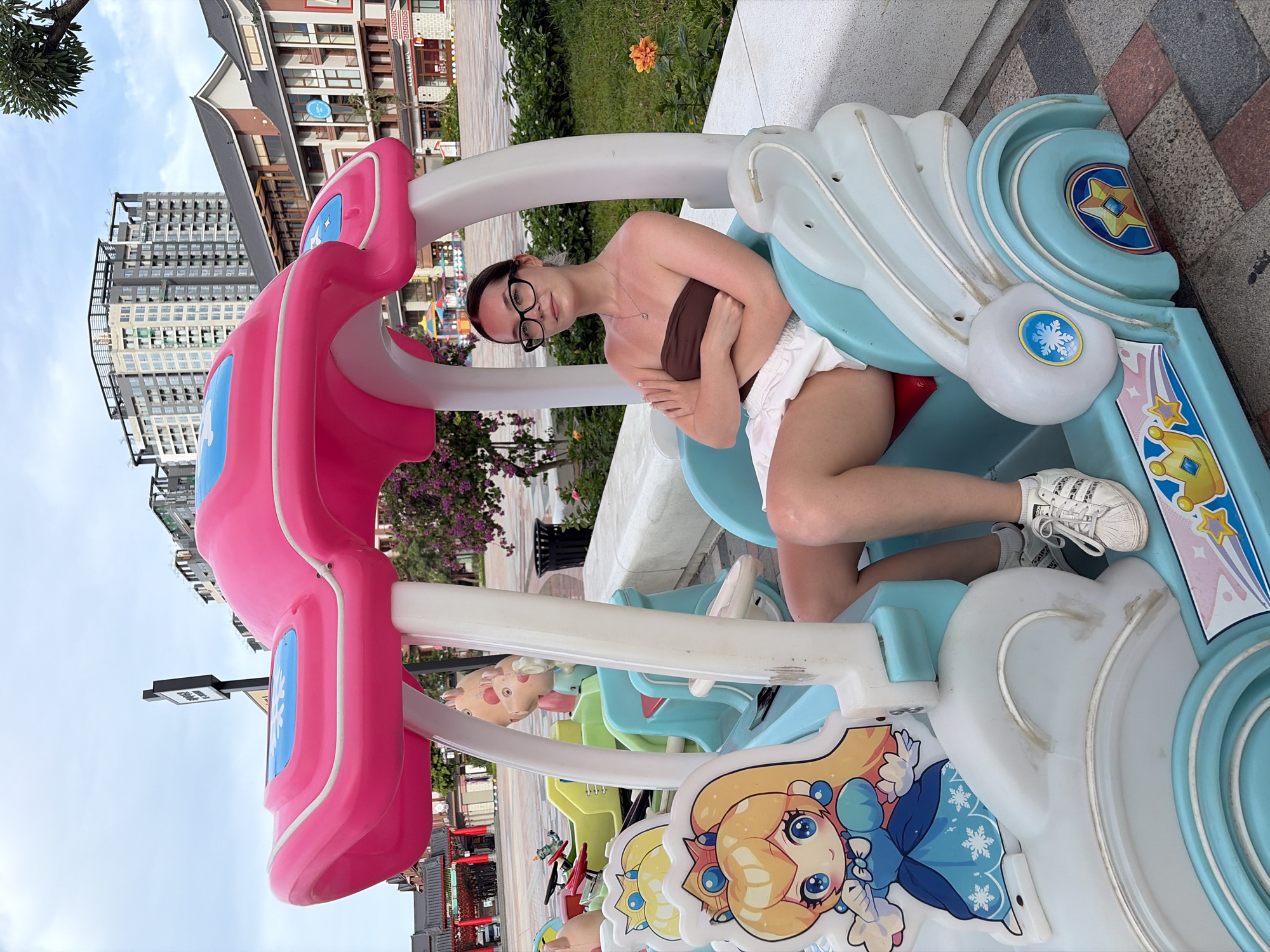 A young woman with dark hair, wearing glasses, a brown strapless top, and white shorts sits in a colorful children's amusement ride shaped like a car with a pink canopy. She has her arms crossed. The ride features a cartoon character, Princess Peach, on the left side. The background shows a street scene with buildings and trees. This scene is located in Nghĩa Trụ Commune, Vietnam.
