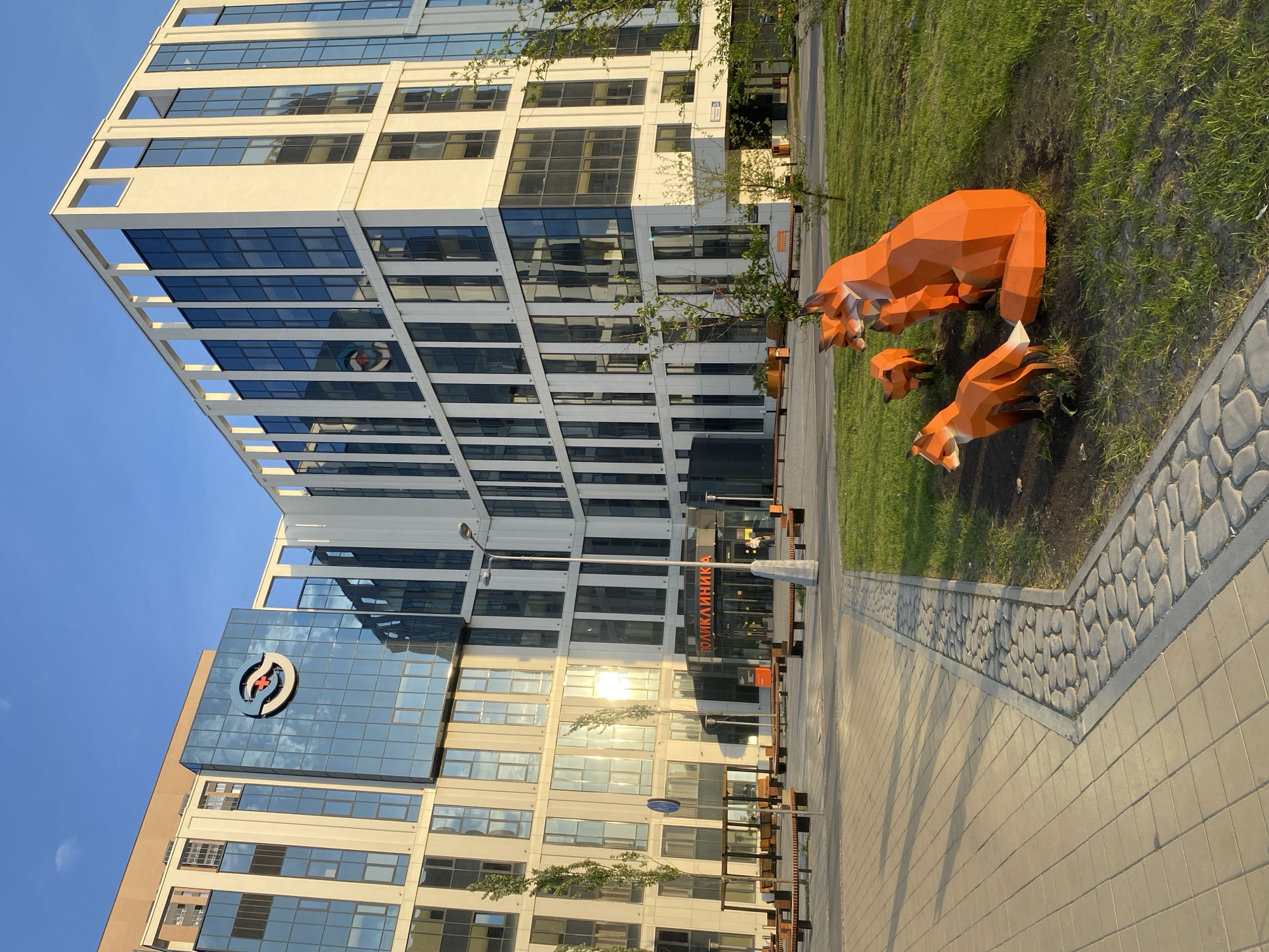 The image depicts a modern urban scene in Yekaterinburg, Russia, featuring a contemporary building and a fox sculpture installation.
**Environment:** The scene is set on a clear day with a blue sky. The area consists of a paved walkway and a grassy patch where the fox sculptures are located.
**Building:** A multi-story building dominates the background. It features a combination of glass and white-colored panels, creating a modern architectural design. The building has a sign in Russian which translates to "Polyclinic".
**Sculptures:** The foreground showcases three bright orange fox sculptures made of geometric shapes. They are situated on the grass.
**Details:** The walkway is constructed with square paving stones and has a border of rounded stones. There are benches near the base of the building. A logo is visible on the side of the building.
**Location:** This location is in Yekaterinburg, Russia.