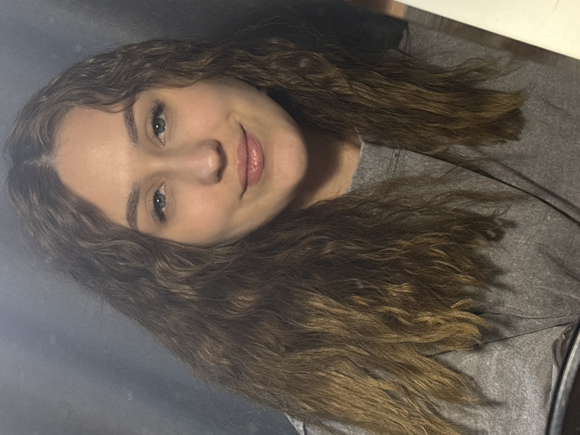 A young woman with long, wavy brown hair is smiling at the camera. She has light skin, green eyes, and pink lips. She is wearing a gray shirt. The background is a mix of gray and white, suggesting an indoor setting. The location in the city of Марковский, Russia cannot be determined from the provided image.