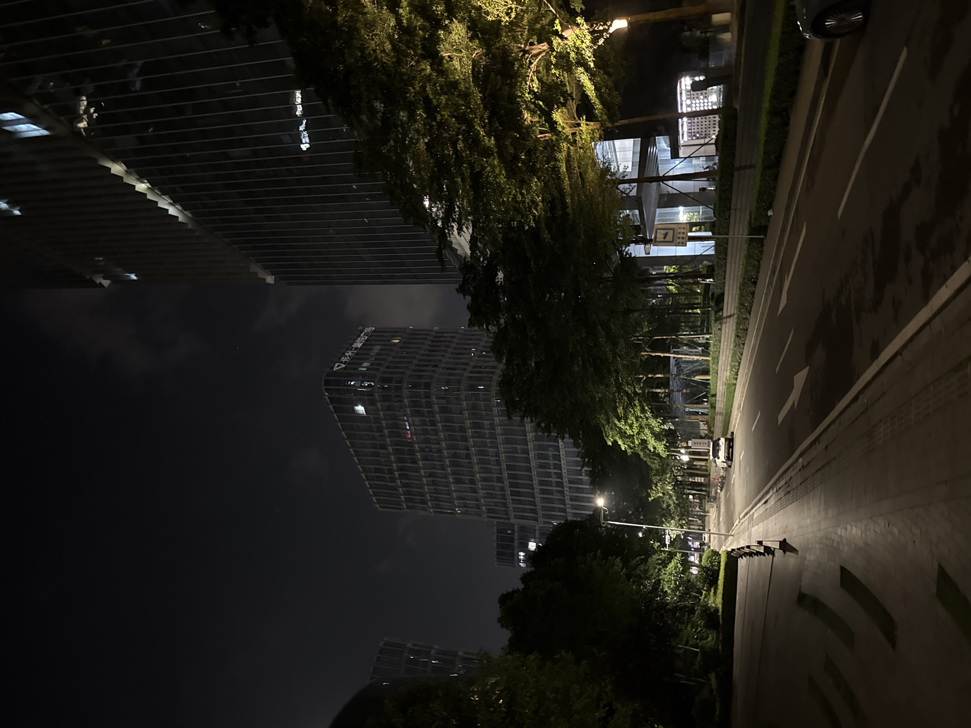This media file depicts a nighttime scene on a city street, likely in the Haizhu District of China. The primary subjects are modern, illuminated buildings and lush green trees. The architecture is characterized by sleek, angular designs with numerous windows reflecting the artificial light. One prominent building displays Chinese characters, appearing to be a company name or advertisement.
The scene is set at night, evidenced by the darkness of the sky and the artificial illumination emanating from the buildings and streetlights. The street itself is paved, with visible lane markings and a pedestrian walkway bordered by greenery. A single car is visible on the road, and a lamppost with a bright light stands tall. The trees are dense and appear to be well-maintained, casting shadows and contributing to the overall atmosphere.
There are no people visibly present in the scene. The overall mood is one of urban tranquility and modern development. The lighting suggests a clear night. The presence of Chinese text on the building is a key location cue, confirming the setting in China. The image is oriented with the buildings on the left and the street on the right, giving a sense of depth and perspective.
