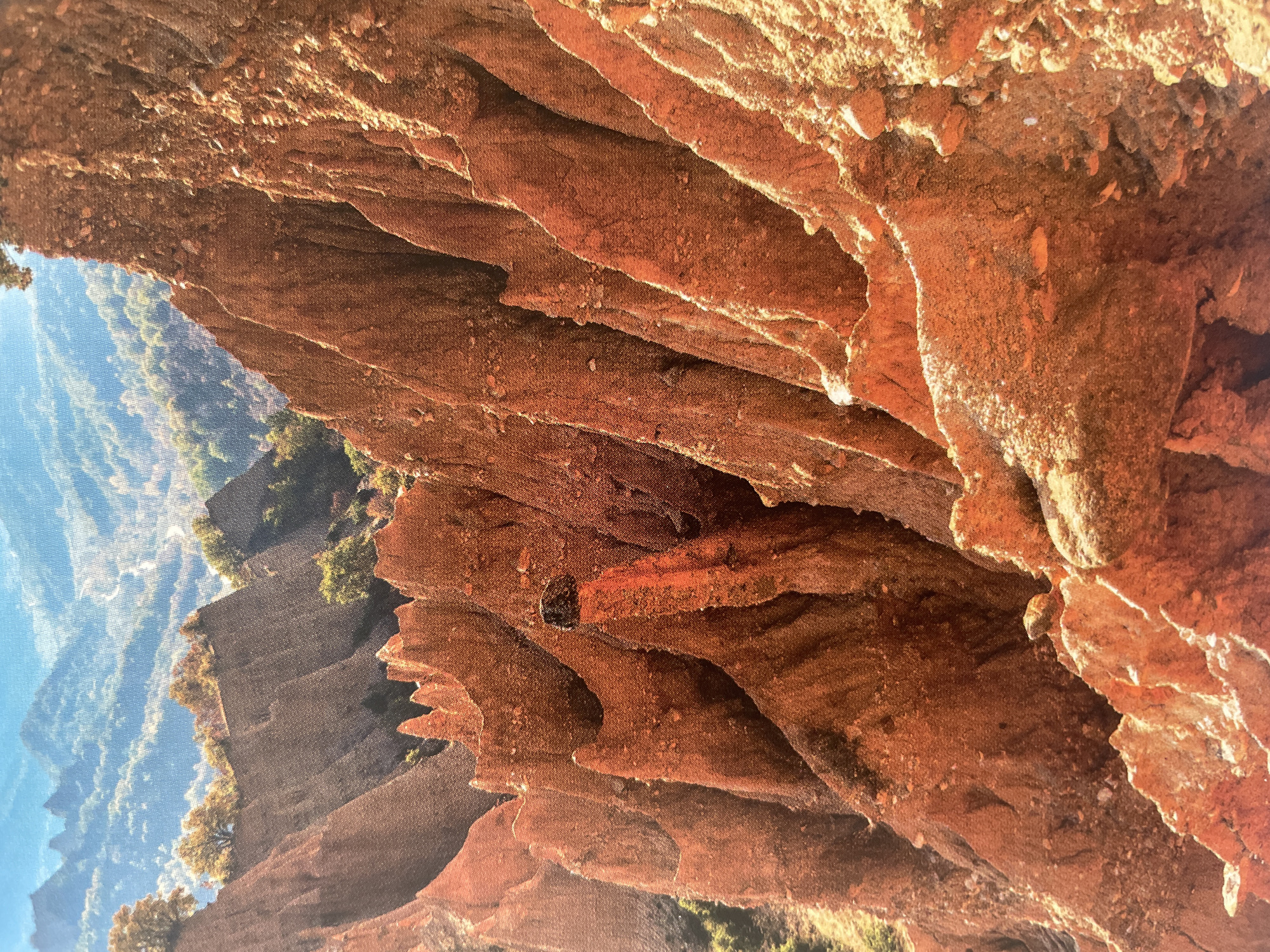The image displays a majestic natural landscape dominated by large, reddish-brown rock formations, likely a canyon or cliff face, under a clear blue sky. The photograph appears to have been taken in a vertical orientation but depicts a horizontal landscape, with the sky on the left and the rock formations filling the majority of the frame on the right.
In the foreground and mid-ground, intricate layers of rock are visible, characterized by their warm, earthy tones of red, orange, and brown. These formations show signs of significant erosion, with jagged edges, deep crevices, and a textured, granular surface. Sunlight, likely from a late afternoon or early morning sun, casts strong highlights and deep shadows, emphasizing the dramatic contours and depth of the rock face. A small, dark, roughly rectangular opening or cavity is visible embedded within the rock in the mid-distance, possibly a small cave entrance or a natural hollow.
In the background, on the opposite side of the canyon or valley, distant mountains or hills are covered with lush green vegetation, appearing hazy and soft against the light blue sky. Scattered green bushes or trees also cling to parts of the rock formations in the mid-ground.
There are no people, animals, or visible man-made objects in the scene, nor is there any visible text. The image captures a moment of serene natural beauty and geological grandeur. Given the context, this striking landscape is located near Sofia, Bulgaria, showcasing the diverse geological features of the region. The weather is clear and sunny, contributing to the vibrant color palette and strong contrasts.