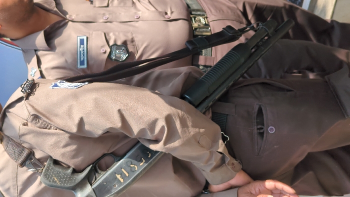 The image displays a close-up of a person wearing a light brown or khaki-colored police uniform. The uniform consists of a short-sleeved shirt with epaulets and pockets, and darker brown pants. On the left side of the uniform shirt, a rectangular name tag is visible, bearing what appears to be Thai script. Below the name tag, a silver-colored badge or emblem is attached to the shirt pocket flap. Another badge, with a blue and white design, is visible on the wearer's left shoulder, near the epaulet.
A black firearm, resembling a pump-action shotgun, is prominently positioned across the torso of the uniformed person. It is secured by a black sling that goes over the wearer's right shoulder and across their chest. The barrel and pump mechanism of the shotgun are visible on the upper left, while the stock and grip are seen towards the bottom right. White markings, possibly numbers or letters like "T13" or "T15", are faintly visible on the side of the shotgun's stock.
The uniformed person's left arm is bent, with the forearm resting across their body, partly obscuring the firearm's sling. Another person's dark-skinned hand is visible in the bottom right corner of the frame, resting near the uniformed person's hip or leg.
The lighting suggests it is daytime, with bright sunlight highlighting parts of the uniform and the firearm. The overall scene captures a moment of a police officer, possibly on duty, with their weapon.