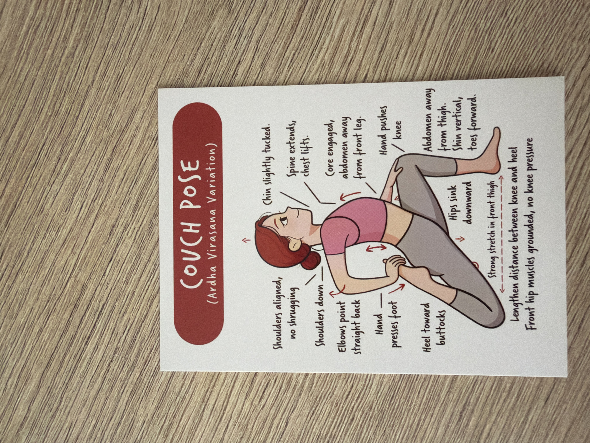 The image is a close-up, overhead shot of a postcard depicting a yoga pose called "Couch Pose (Ardha Virasana Variation)". The postcard is placed on a wooden surface, likely a table, with visible wood grain. The postcard itself is white with a red rounded banner at the top containing the pose name in white text. The illustration shows a cartoon woman in a yoga pose with various anatomical cues labeled in black text. These labels describe the alignment of shoulders, elbows, hands, hips, and legs, as well as the posture of the head and spine. The wooden surface has diagonal lines indicating the grain. The postcard is positioned slightly angled on the surface. There is no information in the image or its context that relates to Pazardzhik, Bulgaria.
