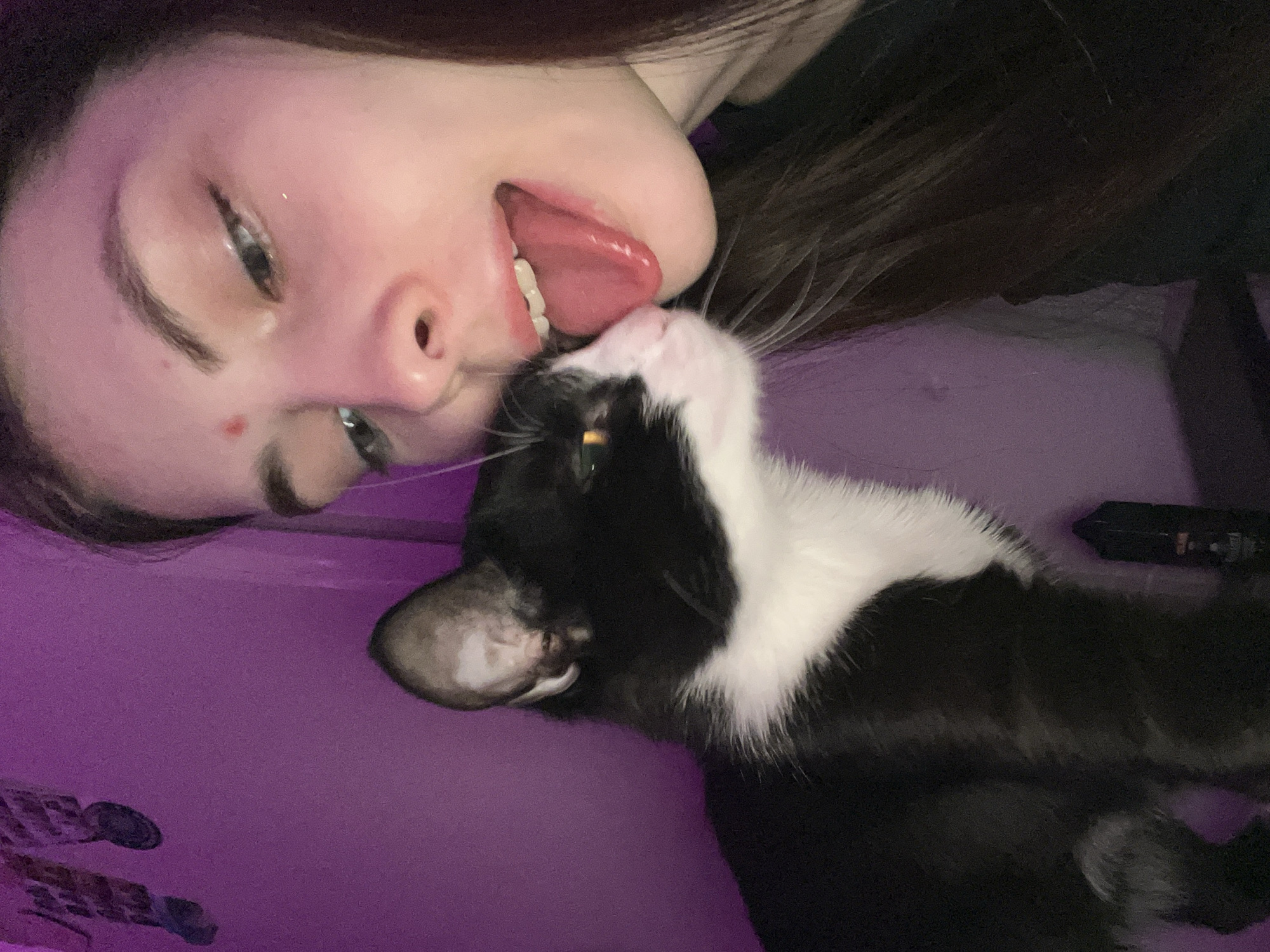 A fair-skinned person with dark hair is lying down, with their head tilted back and their tongue sticking out towards a black and white cat. The cat is positioned close to the person's face. The background is a solid purple color. The person's features include noticeable eyebrows, an eye looking to the side, a nose, and a mole on the cheek. The cat's face is partially visible. The image appears to be taken indoors. The location is not discernable from this image.