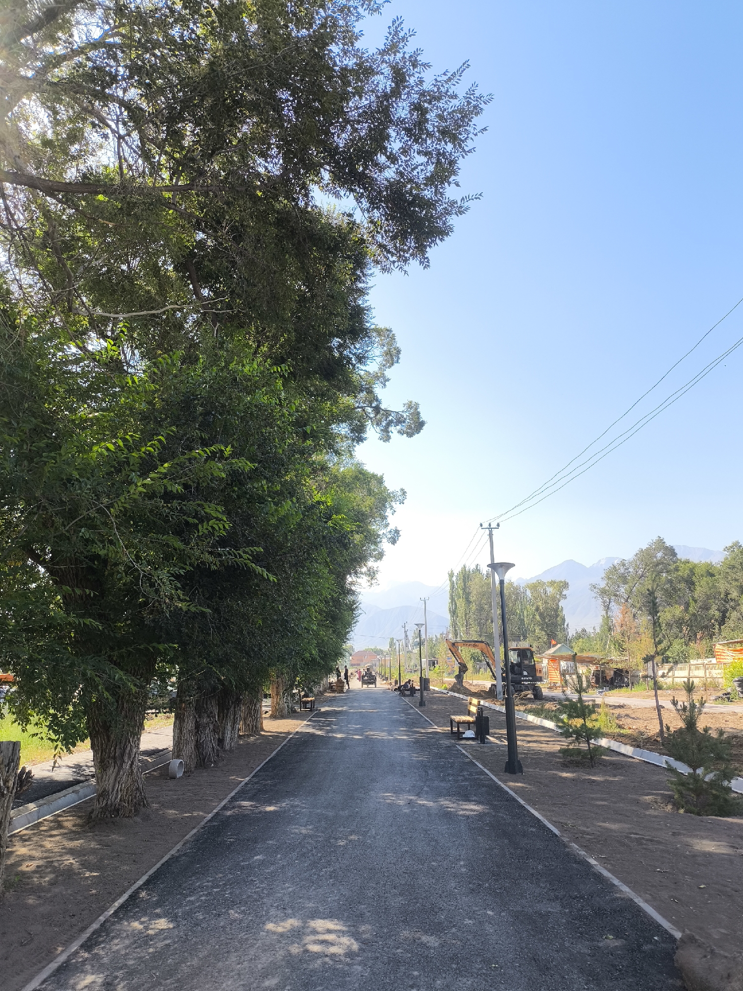 This image depicts a paved pathway in Kochkor, Kyrgyzstan, lined with mature trees on the left and a construction area on the right. The pathway is illuminated by sunlight filtering through the dense foliage of the trees. Several benches and lampposts are visible along the pathway, suggesting it is a public space. In the background, a clear blue sky stretches above a range of distant mountains. The construction area on the right side of the pathway features an excavator, piles of dirt, and what appear to be building materials, indicating ongoing development or renovation. A few people are visible in the distance, walking along the pathway, and a horse-drawn cart is also present. The overall scene suggests a serene, park-like environment undergoing modernization.