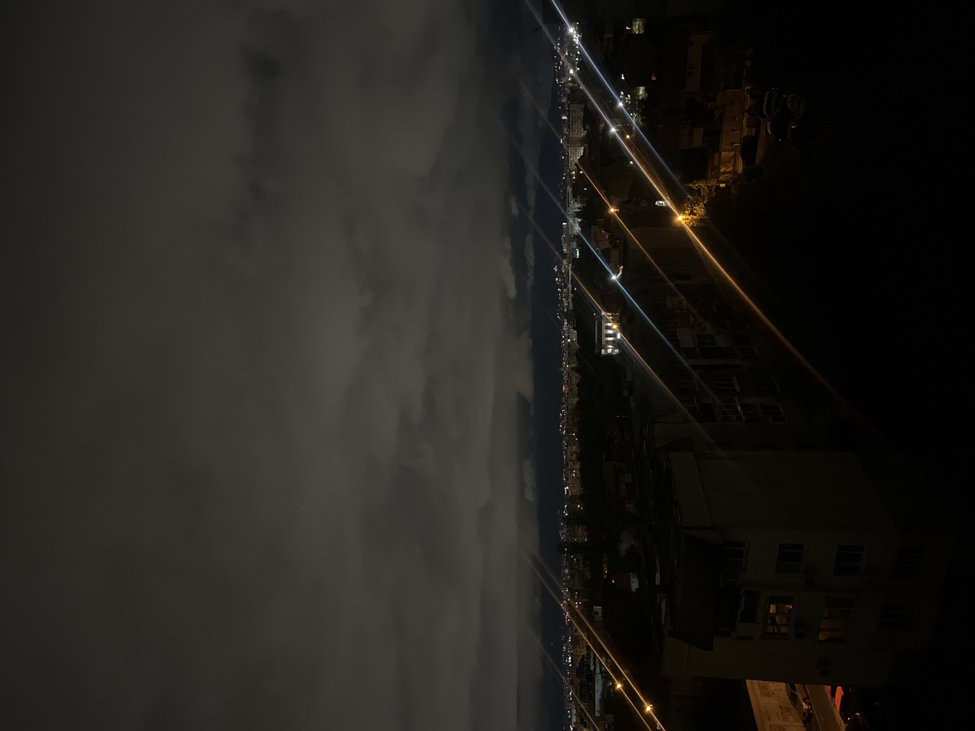 The image presents a panoramic nighttime view of an urban landscape, likely Sofia, Bulgaria, captured from an elevated vantage point. The scene is dominated by a dark, cloudy sky that occupies the upper half of the frame. Below the horizon, a long stretch of the city is visible, illuminated by numerous artificial lights.
The primary subjects are the extensive cloud cover overhead and the distant cityscape. The urban area is characterized by a linear arrangement of buildings, whose presence is mainly suggested by the numerous light sources. These lights appear as distinct, elongated streaks and points of white, yellow, and orange, indicating streetlights or similar fixtures with visible diffraction spikes due to the camera's optics. They line what appears to be a major road or boulevard receding into the distance. In the bottom right foreground, a large, light-colored building is partially visible in deep shadow, with several windows emitting light, revealing hints of its interior. Dark, indistinct shapes scattered throughout the urban sprawl suggest additional buildings or vegetation.
No specific activity or event is taking place; the image captures a static nocturnal scene of the city.
Notable details include the clear indication of nighttime due to the dark sky and reliance on artificial illumination. The weather is overcast, with dense clouds covering the sky. While no specific landmarks are visible to confirm Sofia, the context provided and the general urban architecture are consistent with a European city. The image evokes a quiet, expansive atmosphere characteristic of an urban night vista. There is no visible text in the image.