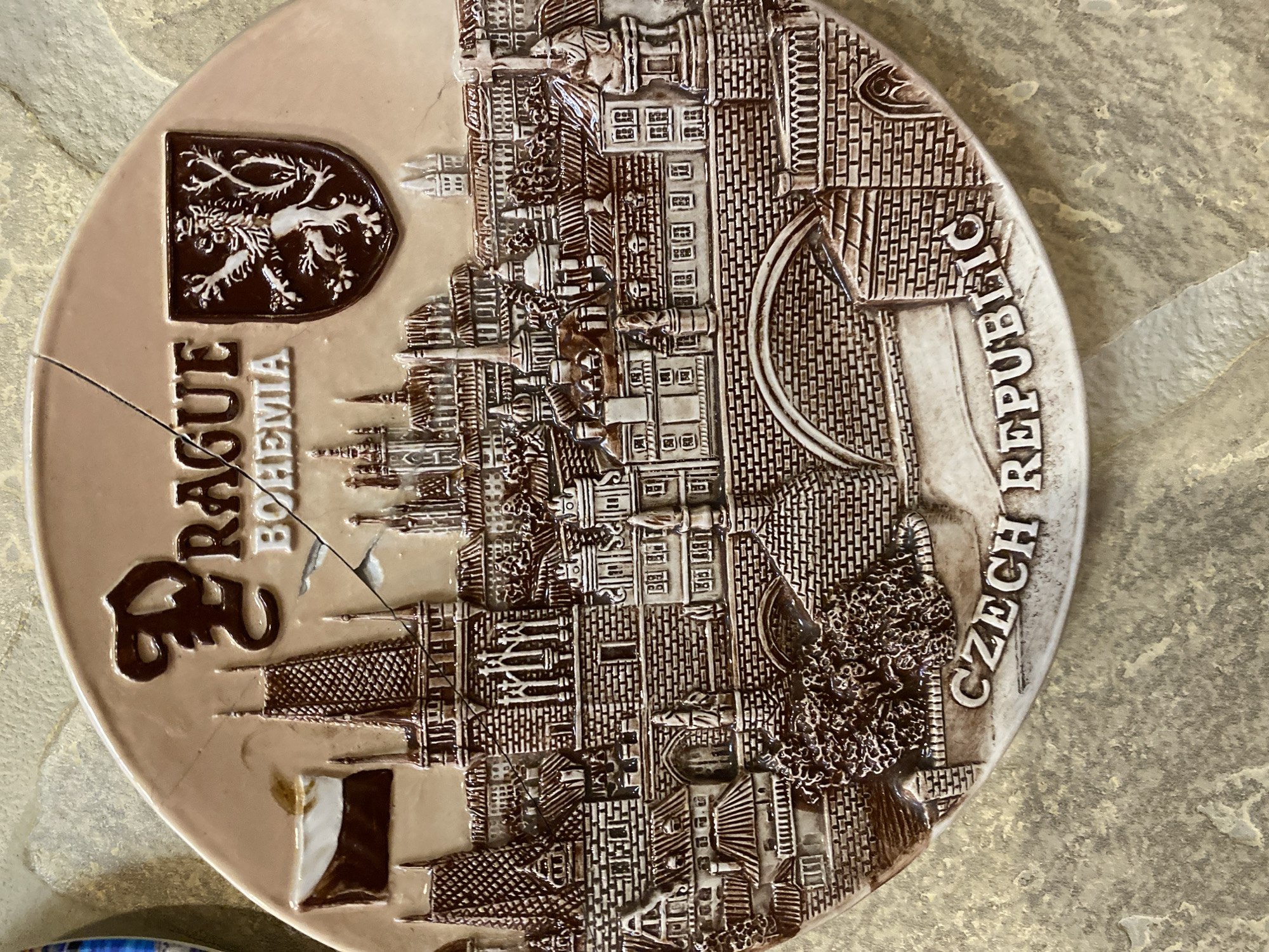 This is a close-up, top-down, slightly angled shot of a cracked, decorative plate. The plate is round and made of a beige ceramic material with a raised, brown relief design. The design depicts a cityscape with intricate architectural details, including brickwork, windows, and arched doorways, likely representing Prague. A stylized crest with a lion or similar animal is also visible on the left side of the plate. The plate is cracked in several places, with visible fragments missing and some discoloration. Text is visible on the plate, spelling out "PRAGUE BOHEMIA" on the left and "CZECH REPUBLIC" on the right, oriented along the curve of the plate. The background appears to be a textured, light-colored surface, possibly a table or countertop, with a mottled pattern. The lighting is consistent and does not suggest a specific time of day or weather conditions. No people or specific activities are depicted; the focus is solely on the object itself. The setting's location is given as Novaya Usman', Russia, but this is not visually evident from the image.