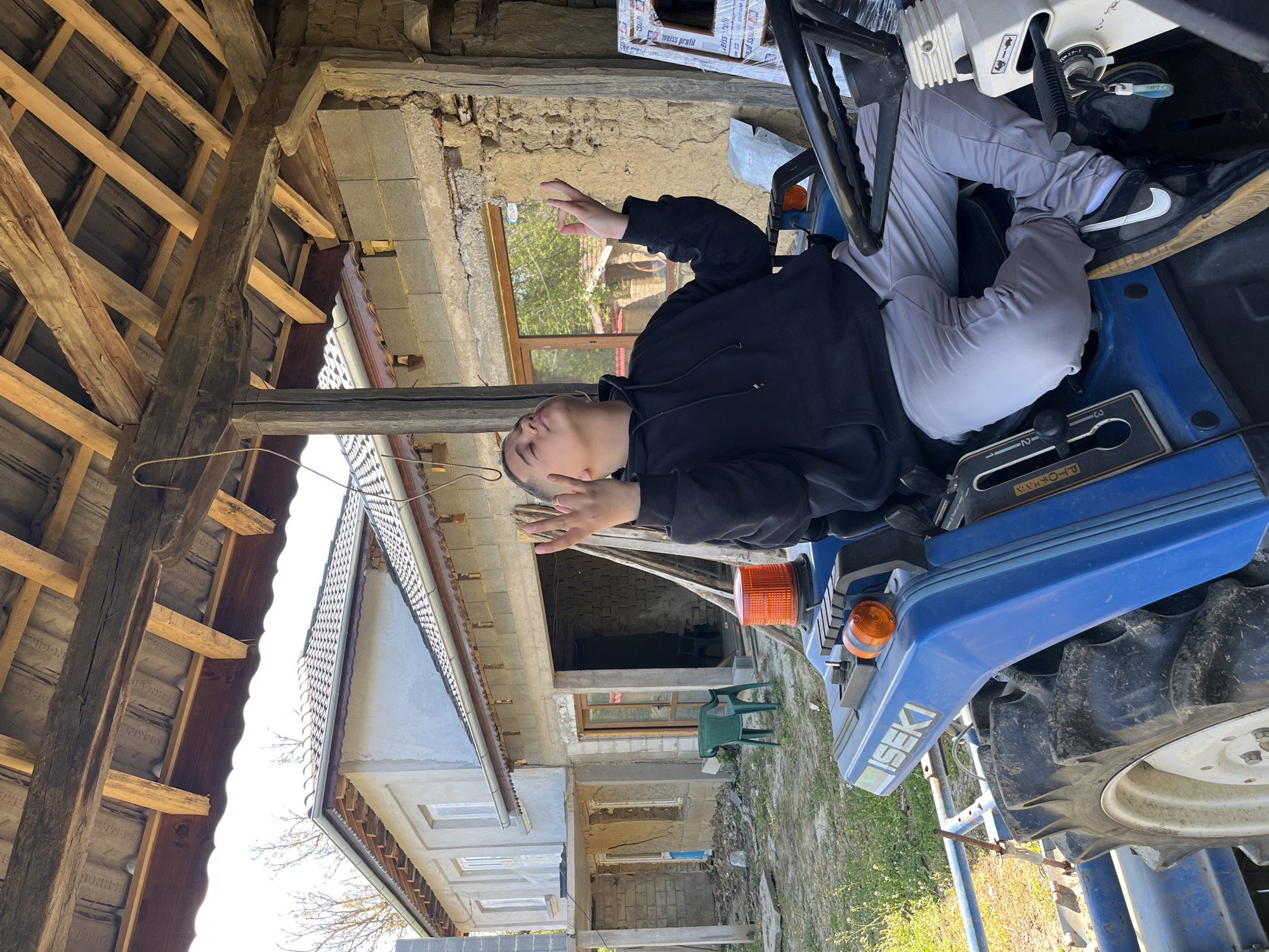 The image captures a person seated on a blue ISEKI tractor, likely in a rural setting. The person is wearing a black hoodie and light grey pants. The background features a partially renovated or under-construction building, with exposed brick and concrete blocks. There is also a wooden roof structure visible, along with a glimpse of vegetation, indicating an outdoor location.
Based on the text "ДОНЕЕ ОРМАТ," "ДОНЕЕ ОРМАТ NOGIH," and "Weiss profil" which may refer to construction materials and the presence of the Cyrillic alphabet, it is plausible that the location could be Rish, Bulgaria, as specified. The construction material manufacturer Weiss Profil operates in Bulgaria. However, without further information or distinct location markers, confirmation is limited.