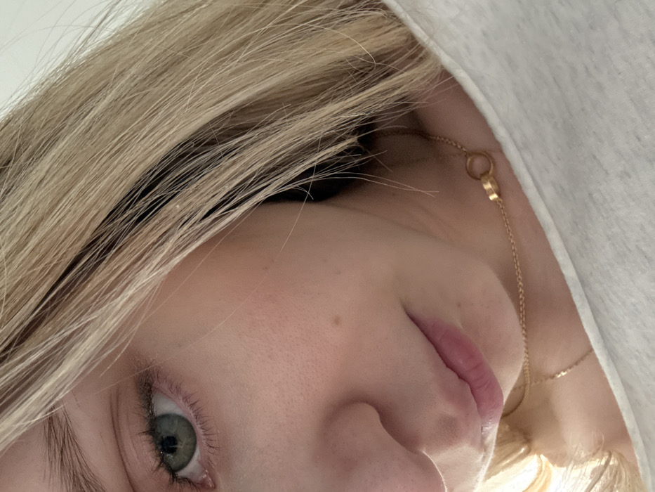 The image depicts a close-up, angled view of an individual in an indoor setting within the city of Coral Gables, United States of America. The frame focuses on the left side of the subject's face, extending to a portion of the neck and upper chest, observed from a slightly elevated vantage point.
The subject has fair skin and light blonde hair, with some darker streaks, covering the right side of the image and partially obscuring the subject's right facial features. The visible left eye exhibits a greenish-grey iris, a dilated pupil, and natural eyelashes, with a faint application of light eyeshadow on the eyelid. The left nostril, the bridge of the nose, and the subject's mouth are partially visible; the lips are naturally pink and slightly parted. A small, dark mole is present on the left cheek.
The subject wears a light-colored, soft-textured garment, likely a t-shirt or hoodie, visible across the lower right section of the frame, showing the neckline and shoulder. A thin, gold-colored chain necklace is present around the neck, featuring a small, circular, gold-colored pendant (appearing as two interlocking rings). A second, finer gold-colored chain is partially visible beneath the primary necklace.
The background is uniformly light-toned and blurred, consistent with an interior environment receiving soft, ambient illumination. No additional subjects, objects, specific actions, or interactions are discernible within the scene.