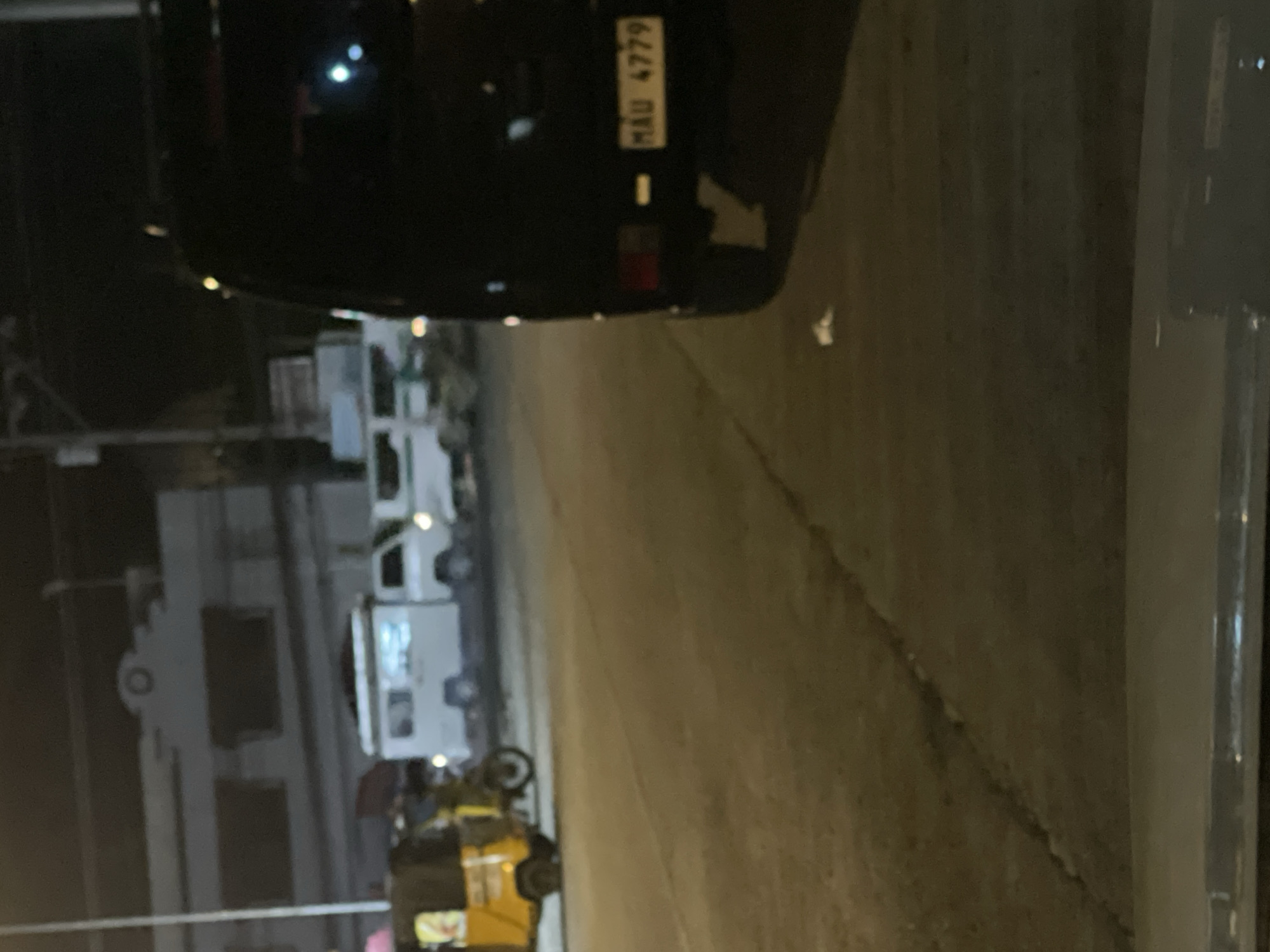 The image captures a street scene at night in Davao City, Philippines. A dark-colored SUV is parked in the foreground on the right, with its license plate reading "MAU 4779". To the left of the SUV, several white vans and a yellow three-wheeled auto-rickshaw are visible. In the background, a light-colored building with dark windows is illuminated. The overall scene suggests a typical urban environment at night, possibly a street or parking area.