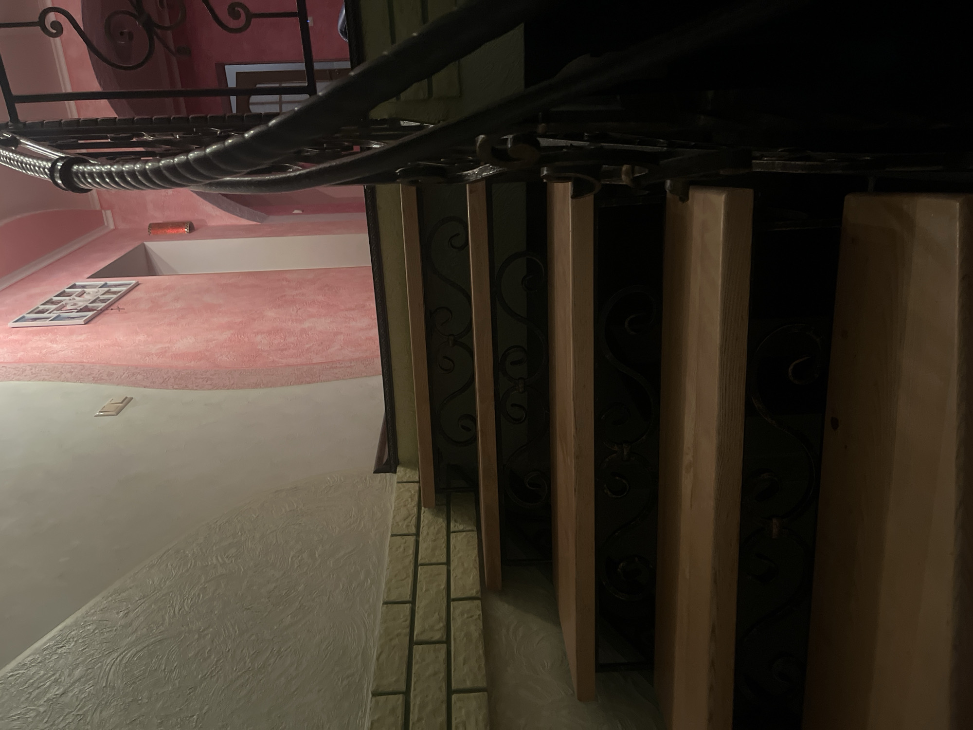 The image shows a wooden staircase with ornate metal railings. The walls are a light color and textured, with a section of what appears to be brickwork visible near the bottom left. The staircase is dimly lit, with shadows obscuring some of the details. A pink wall is visible in the background, along with a framed picture and a red light fixture. The scene appears to be indoors, likely in a residential building or a public space. There are no people visible in the image. The overall impression is one of a dimly lit, somewhat eclectic interior space.