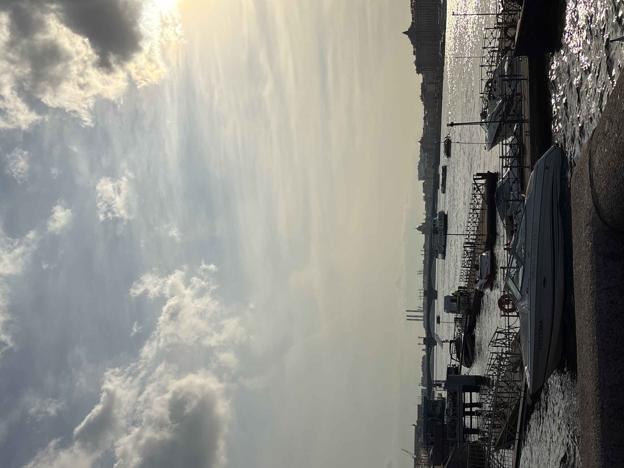 The image depicts a waterway with several boats docked along a stone embankment, presumably a river or canal. Buildings in the background suggest an urban environment. The presence of boats and architectural style indicates this could be Saint Petersburg, Russia. The angle of the sun suggests it is a daytime view. The sky is filled with clouds, some of which are darkened.
