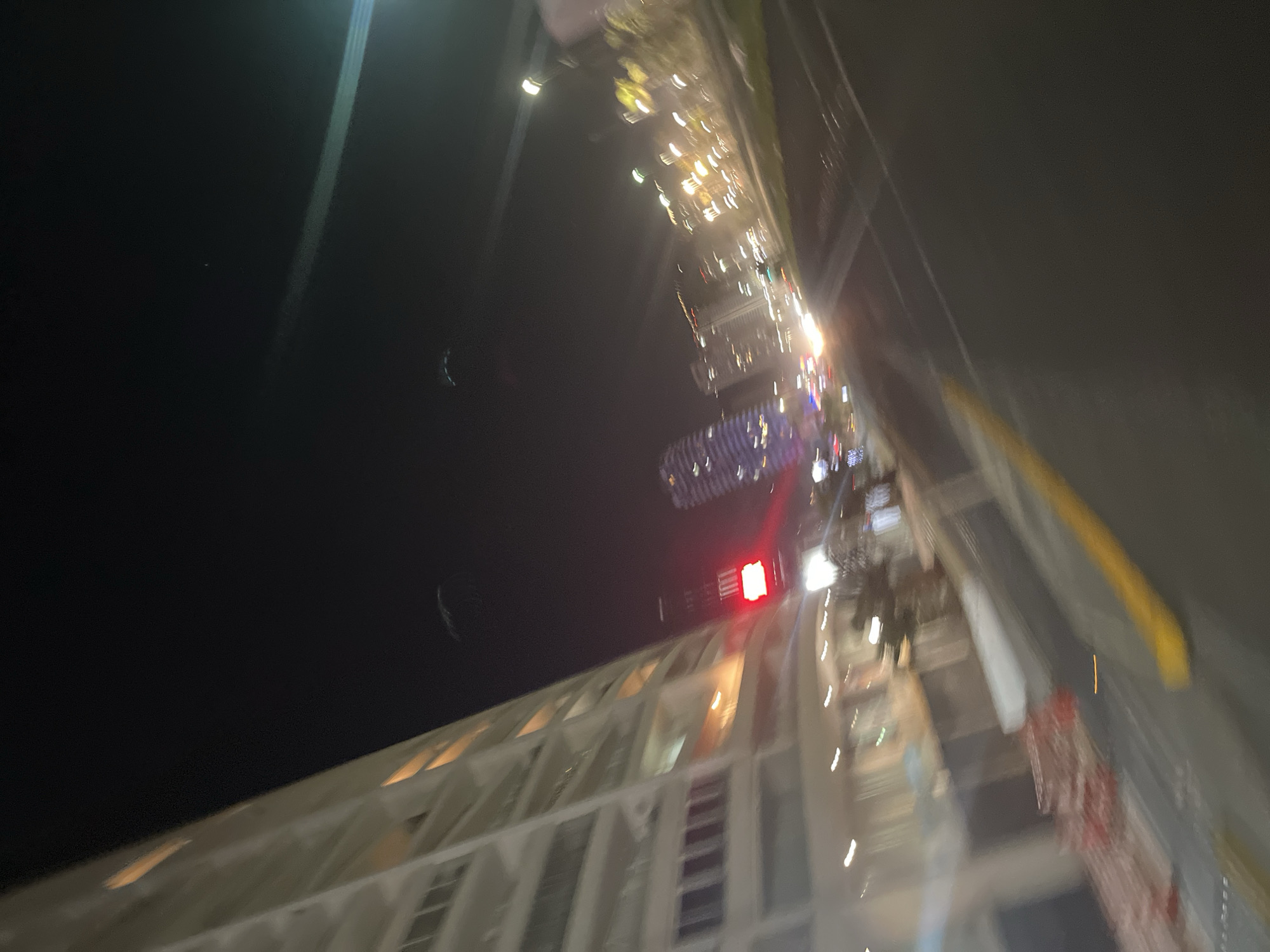 The image depicts a blurred, low-light urban night scene. On the left, a tall, multi-story building with numerous balconies and illuminated windows rises diagonally into the frame. A red traffic light is visible near the base of this building. The street curves from the bottom right towards the center, bordered by a yellow curb or barrier on its left edge. In the mid-ground and background, multiple other tall structures are discernible, some adorned with various colored lights, creating bokeh effects. Streaks of light indicate vehicular traffic moving in the distance. The sky is dark, consistent with nighttime conditions. No individuals or specific actions are clearly identifiable due to the significant motion blur and low resolution. The environment suggests an active city street.