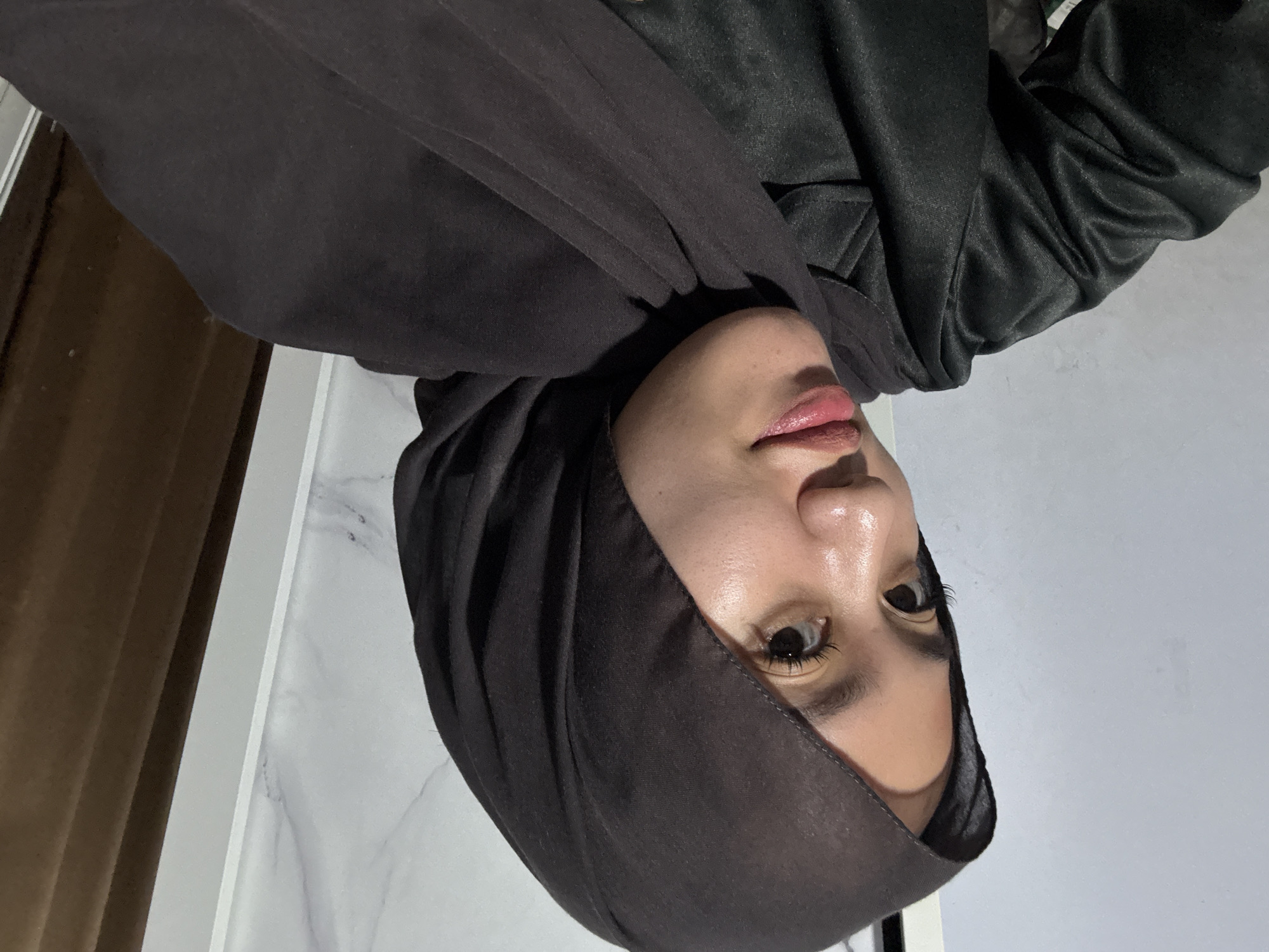A young woman wearing a dark hijab and a shiny black shirt is captured in a close-up selfie. Her face is turned to her left, with her right eye clearly visible and her left eye partially obscured by her hijab. She has on makeup, with a shimmery eyeshadow and pink lipstick. The background shows a light-colored wall with a subtle marble pattern to her right, and a plain light gray wall to her left. There is also a glimpse of a brown curtain on the far right. The lighting suggests an indoor setting, possibly a room in Gadingrejo, Indonesia.