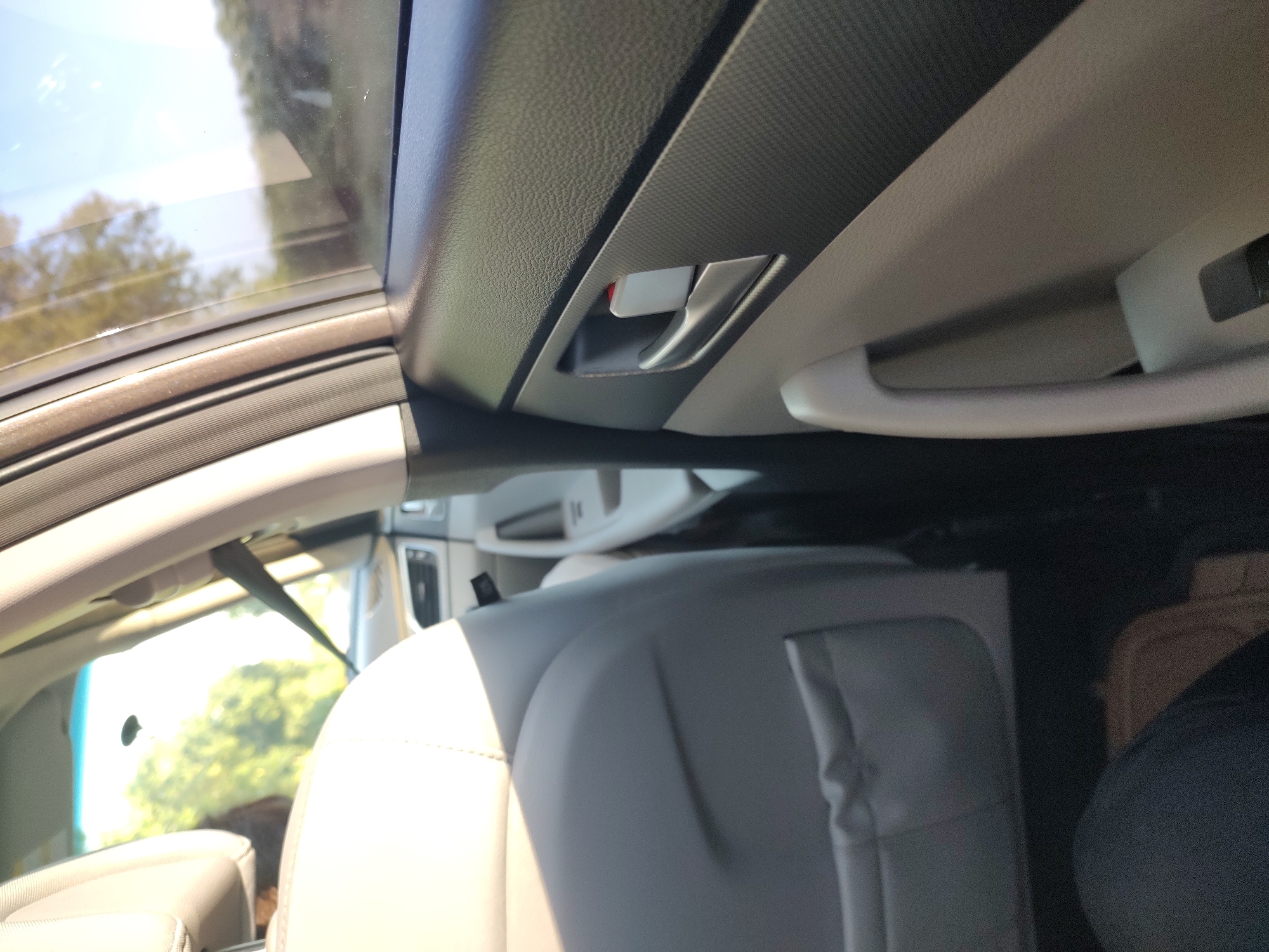 The image shows the interior of a car, likely a van or SUV, with a view from the back seat looking forward and slightly upward. The scene is bathed in bright, natural light, suggesting it's daytime. The weather appears clear, as evidenced by the bright sky visible through the windows.
The primary subjects are the car's interior elements. We see light-colored leather seats in the foreground, with a darker gray seat back behind them. Above the seats, the car's headliner is visible, made of textured, dark gray material. A light-colored grab handle extends across the top of the frame. A red and silver interior door handle or latch is also visible on the dark gray paneling.
Through the windows, we see a blurred background of green trees and a bright blue sky, indicating the car is likely on a road trip or parked in a scenic outdoor location. There are no people visible in the frame. The overall impression is one of a modern, clean car interior.
The image has a slightly tilted perspective, with the car's interior elements dominating the frame. The details suggest a focus on the passenger experience within the vehicle. The location context provided, Karapelit, Bulgaria, is not directly evident from visual cues within the image itself. There is no visible text in the image.