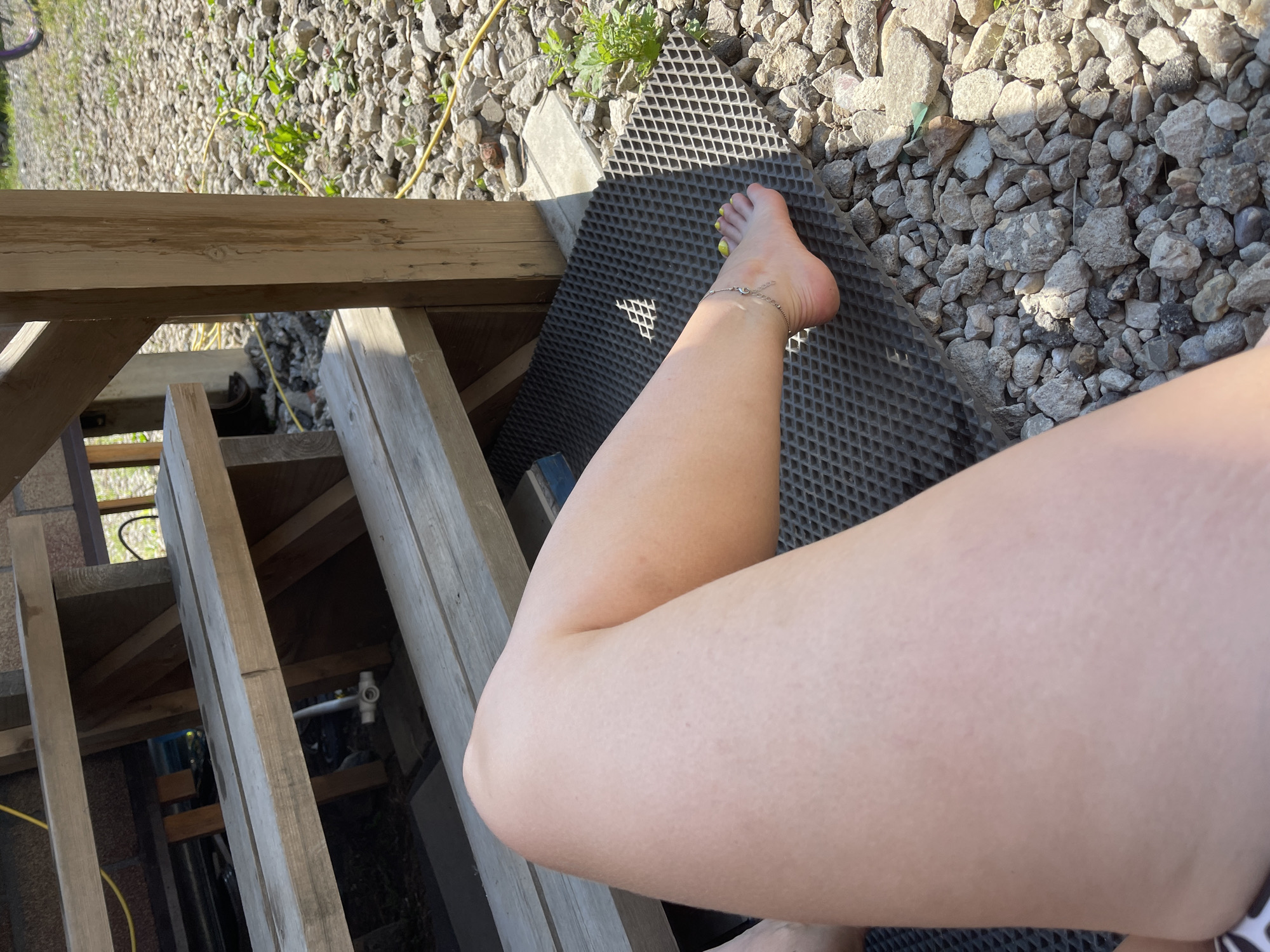 A close-up, high-angle, medium shot shows a person's bare legs and feet resting on a grey textured mat placed on a gravelly ground. The legs are lightly tanned and show minor skin texture. A delicate silver anklet with a small charm is visible on the left ankle. The feet are bare, with toenails painted a bright yellow. Behind the legs, wooden steps are visible, part of what appears to be an outdoor structure. The environment is sunny with shadows cast by the steps. The ground is covered with small grey and tan stones and some sparse green vegetation. The scene is outdoors, likely a backyard or patio area. No city or specific location in Мосметаллоконструкция Онт, Russia is visible or implied by the image itself.