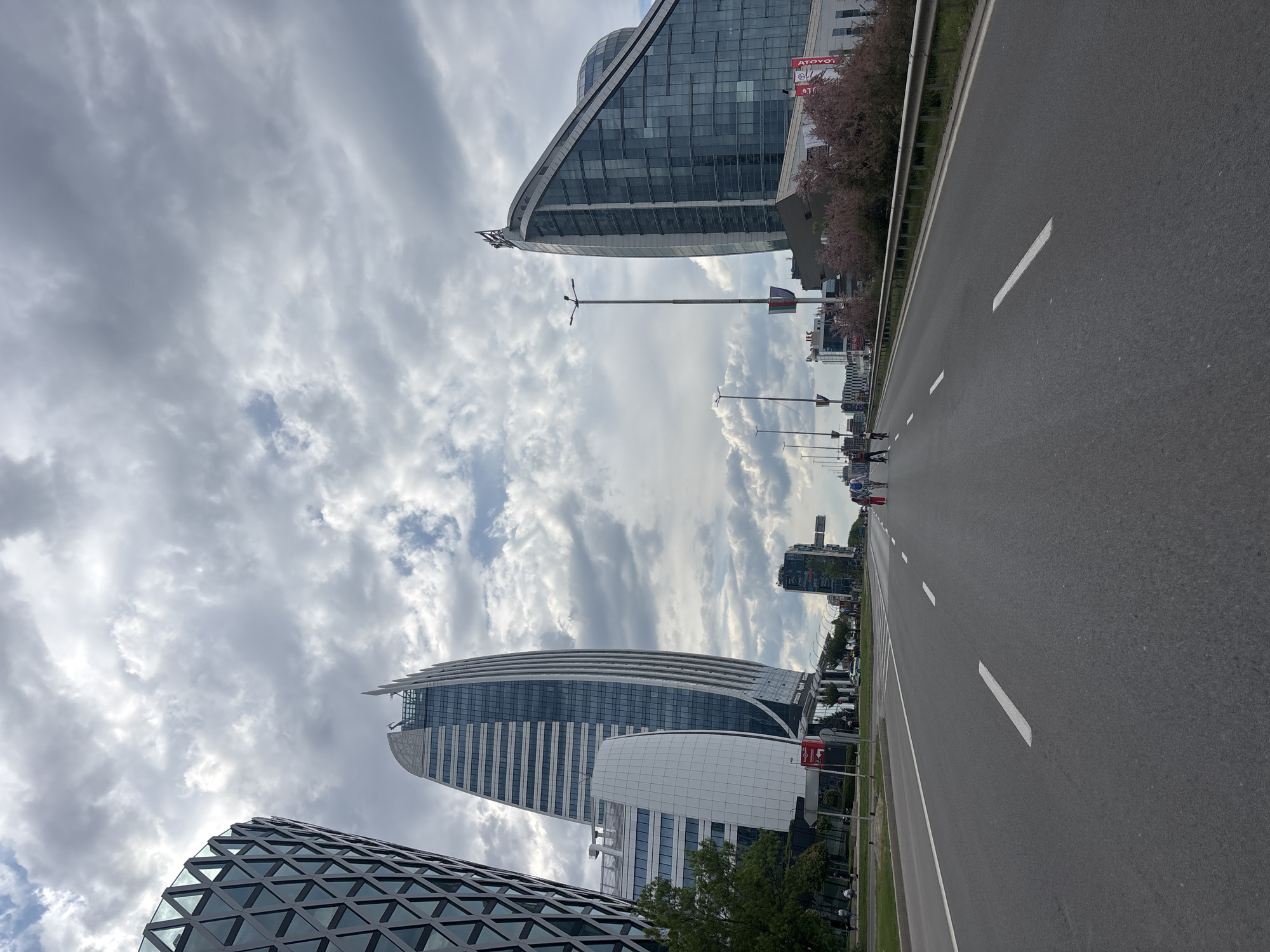 The image shows a street scene in Sofia, Bulgaria. The perspective is from ground level, looking along a wide road with white lane markings.
Several modern buildings of notable design are visible, including one with a distinctive triangle pattern facade. Another prominent building has a curved, elliptical shape. There are also multiple glass and steel office structures.
The sky is filled with dramatic clouds. Pedestrians are visible on the right side of the road. Streetlights are present along the side of the road, with roadside vegetation and advertising.