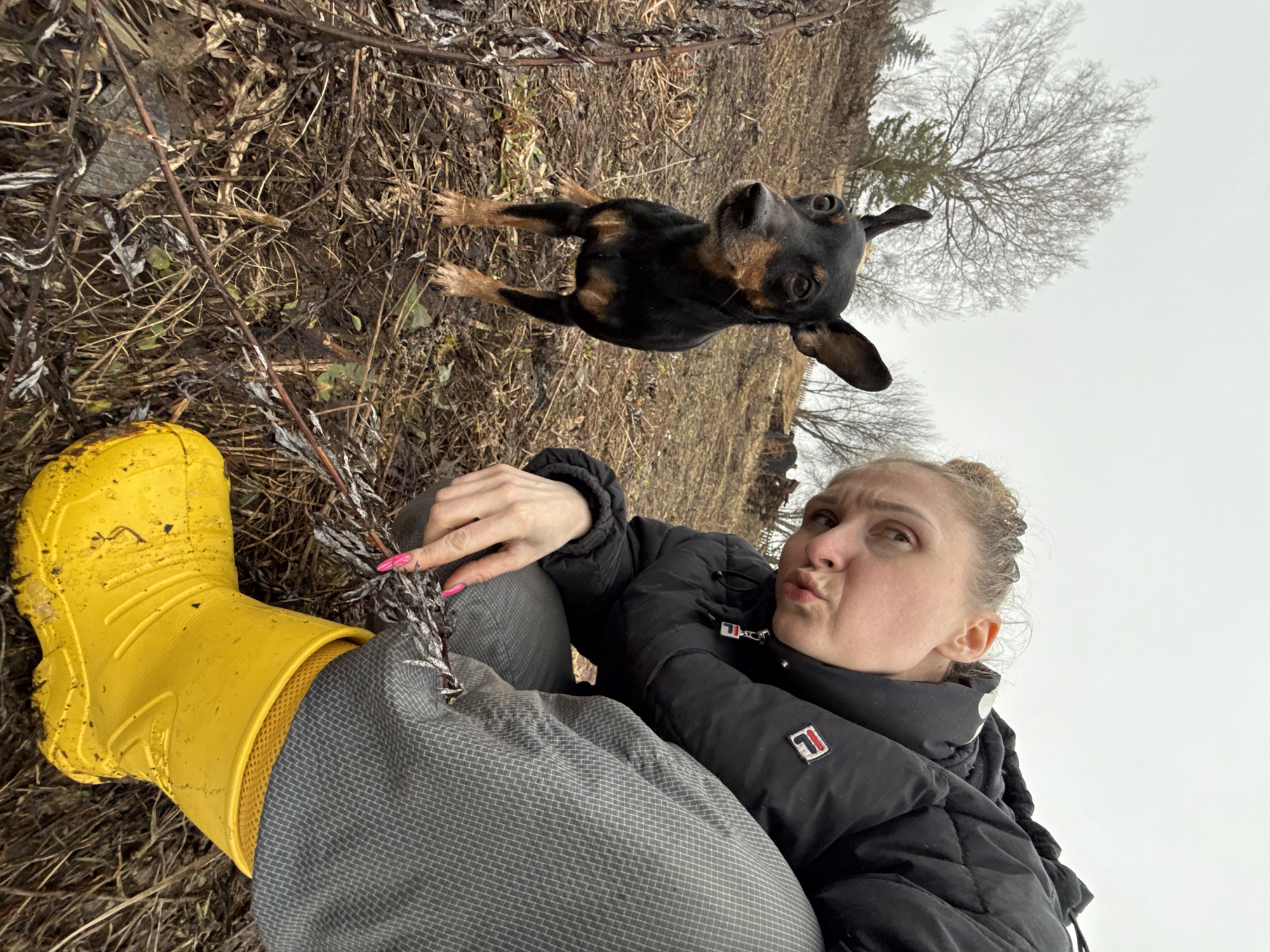 The image appears to be a selfie taken outdoors. A woman is sitting with her legs extended towards the lower left corner. She is wearing grey pants, a black puffy jacket, and a bright yellow rubber boot on her left foot. Her fingernails are painted pink. She is making a pouting face, looking up and to her right.
A small black and tan dog is suspended in the air above her, its body oriented towards the upper left corner. The dog's head is tilted upwards, and its ears are floppy.
The ground is covered in brown foliage, sticks, and dirt. In the background, there is a leafless tree against a grey sky. The location is Nizhnyaya Chermoda, Russia.