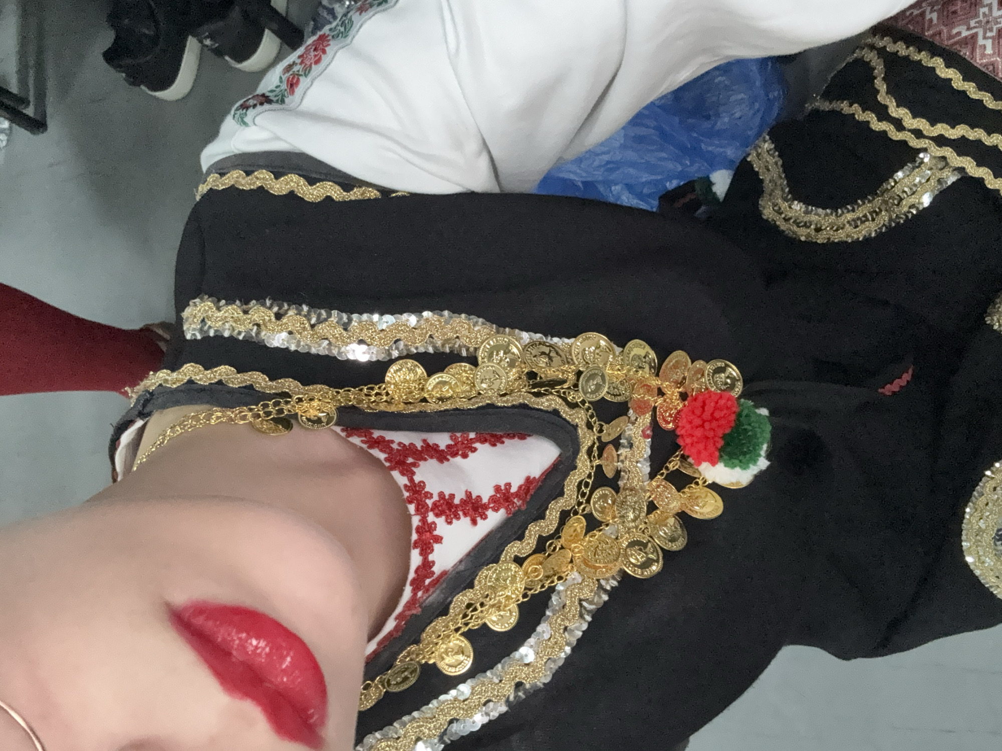 A close-up photograph shows a person wearing traditional clothing. The individual has red lipstick on and is wearing a black garment adorned with gold coins, sequins, and gold braid. Underneath the black garment, a white piece of clothing with red floral embroidery is visible. A decorative element featuring red, white, and green pom-poms is also present. The setting appears to be indoors. The image does not provide enough information to determine the location as Belgrade, Serbia.