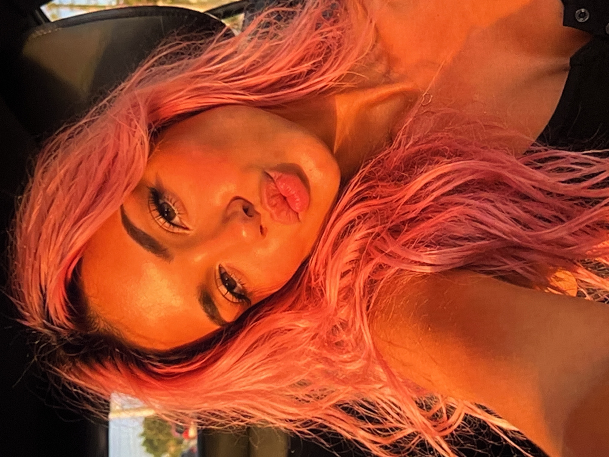 A young woman with vibrant pink hair is pictured in a close-up, selfie-style shot. She is inside a vehicle, as evidenced by the car seat headrest visible behind her. The setting appears to be outdoors during the golden hour, with warm, orange-toned lighting bathing her face and hair. Her eyes are open and looking towards the camera, and her lips are puckered slightly as if for a kiss. She is wearing a dark-colored top, and a delicate necklace is visible around her neck. The background shows a blurred view of trees and what might be buildings, suggesting an urban or suburban environment.
