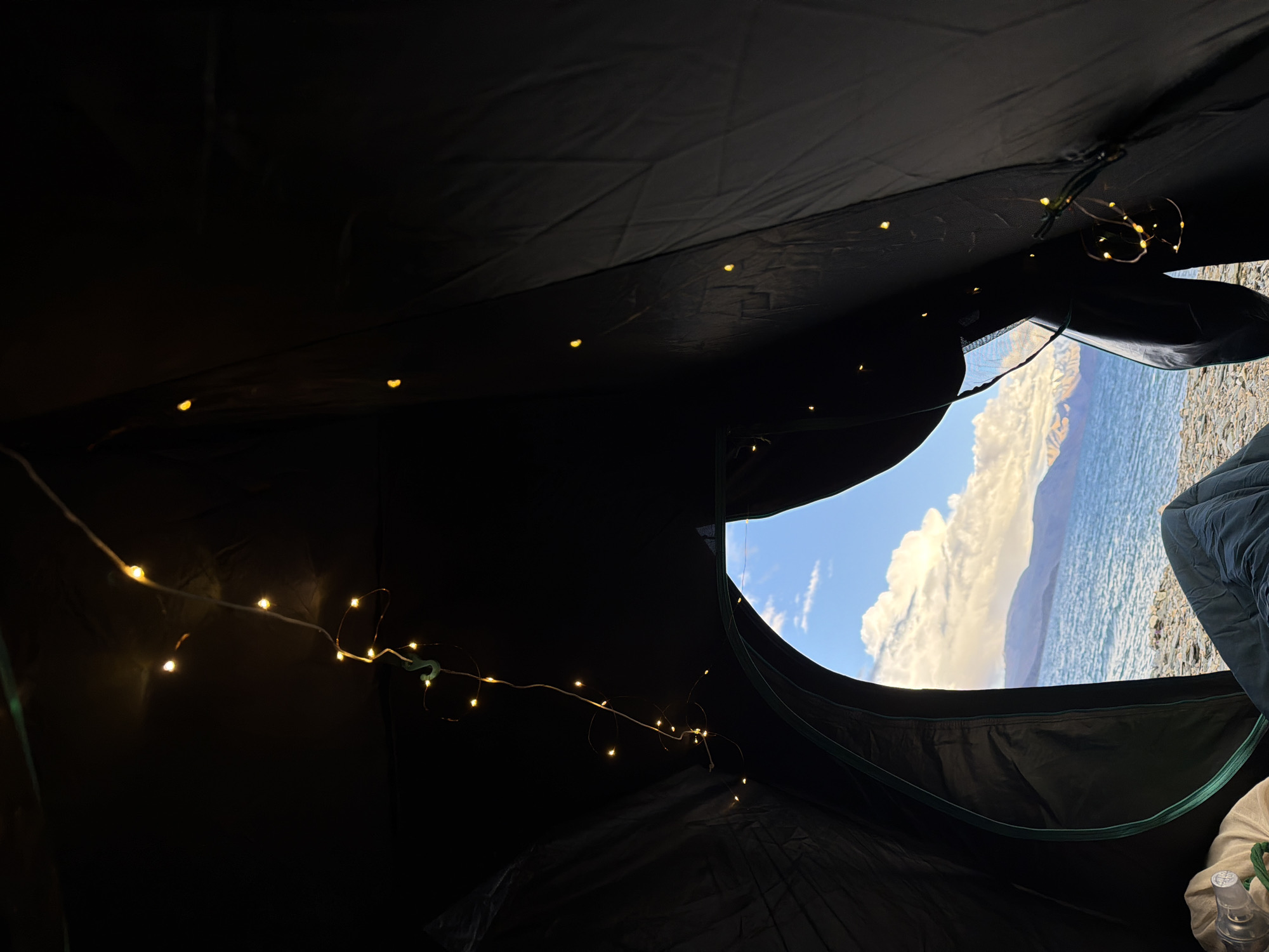 The image shows the interior of a dark tent with a view of a body of water, mountains, and a sky with clouds through the open entrance. A string of small, yellow fairy lights is draped along the inside of the tent. In the foreground, on the left side, a sleeping bag and a white bag are visible. The ground outside the tent appears to be made of pebbles. The location is described as Unknown, India.