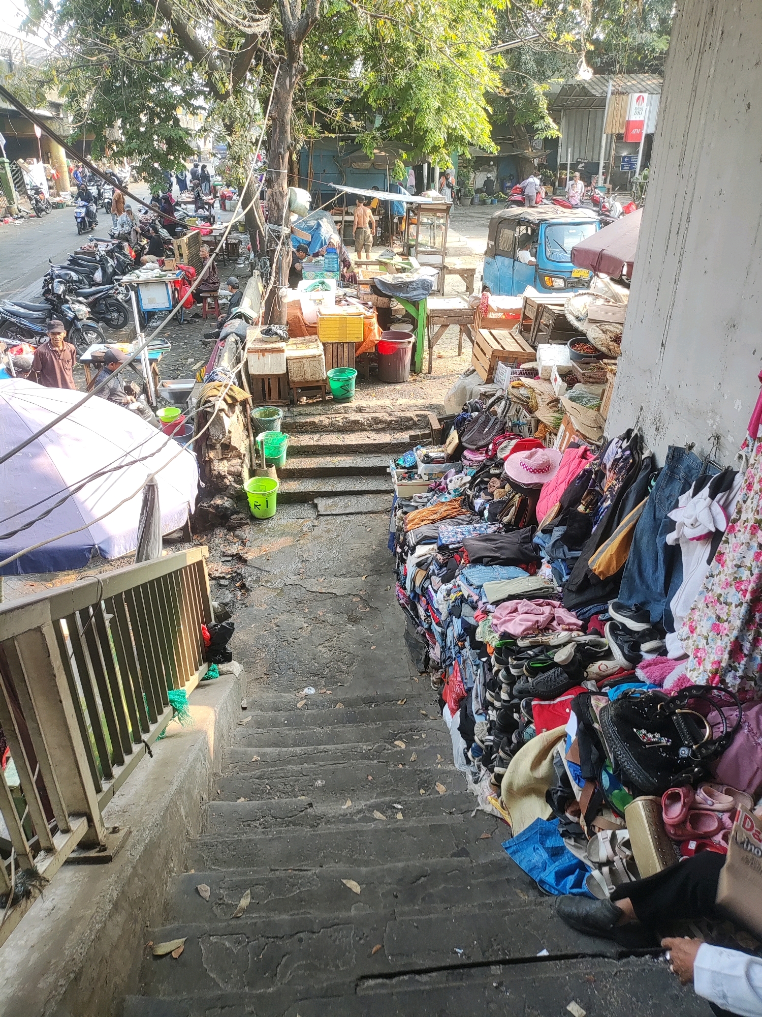 This is a daytime, eye-level shot of a busy street market scene in East Jakarta, Indonesia, potentially near Pasar Rawa Bening or a similar flea market. The foreground features a set of concrete stairs descending into a lower area, with a metal railing on the left. To the right of the stairs, a large pile of clothing and shoes is displayed for sale, including jeans, t-shirts, dresses with floral patterns, sneakers, and sandals. Some bags are also visible in this merchandise pile.
At the top of the stairs, the market extends onto a paved street. On the left side of the street, numerous motorcycles are parked. People are browsing various stalls and interacting with vendors. Several stalls have tables displaying goods, some covered by umbrellas. A blue, three-wheeled vehicle (likely an 'Bemo' or similar public transport) is parked on the street. Trees with dense foliage provide shade over parts of the scene. The overall atmosphere suggests a vibrant, informal marketplace.