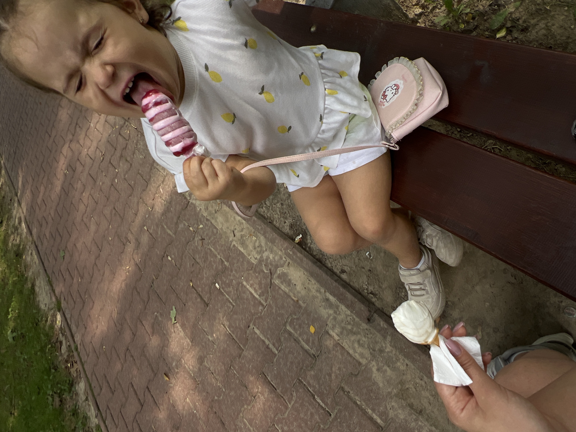 A young girl with her mouth open is eating a pink and white ice pop while sitting on a park bench. She is wearing a white t-shirt with yellow lemon patterns, white shorts, and beige sneakers. A small pink purse with a Hello Kitty design rests on the bench next to her. In the foreground, a hand holds out a white napkin to a partially visible ice cream cone. The girl is sitting on a dark brown wooden bench in what appears to be a park with a red brick path behind her. The scene is set in Lublin, Poland.