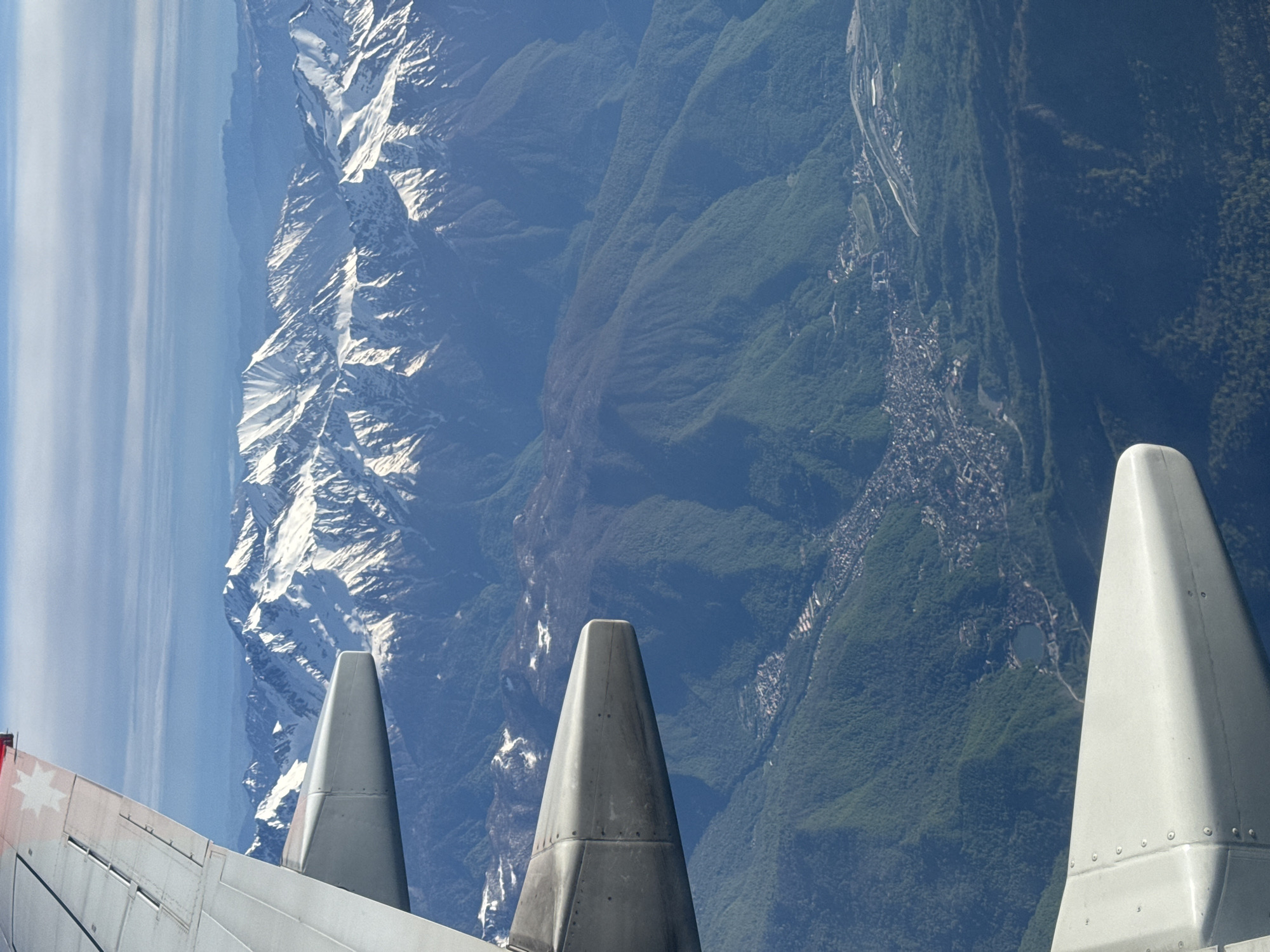A landscape shot from an aircraft shows snow-capped mountains, a valley with a town nestled in it, and the wing of the aircraft with three vertical stabilizers. The snow-covered mountain range is on the left, and next to it is a series of forested mountains. A town is visible in the valley between the mountains, with buildings and roads snaking along the terrain. The wing of the aircraft is in the foreground, taking up the lower portion of the frame. The vertical stabilizers are gray and metallic, contrasting with the natural scenery behind them. The location of Sochi, Russia, is confirmed through the geographic features of the Caucasus Mountains.