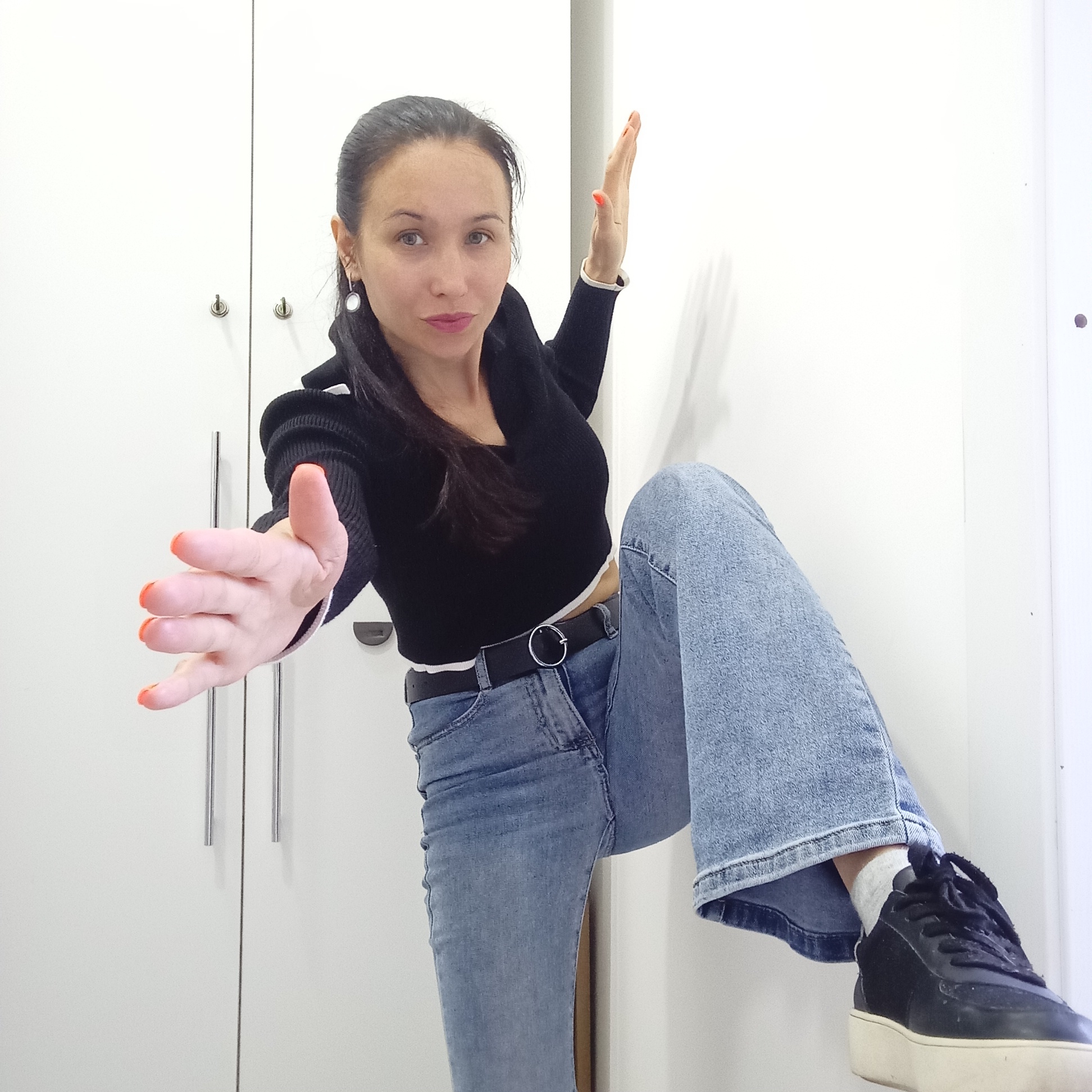 A woman poses indoors against a backdrop of white cabinets. She wears a black cropped sweater over a white top, blue jeans with a black belt, and black sneakers with white soles, visible on her raised leg. Her hair is pulled back, and she has an earring in her left ear. One arm is extended towards the camera, while the other is raised beside her. Her nails are painted orange. The image appears to have been taken at a close range. There is no information to verify that the picture was taken in Izhevsk, Russia.