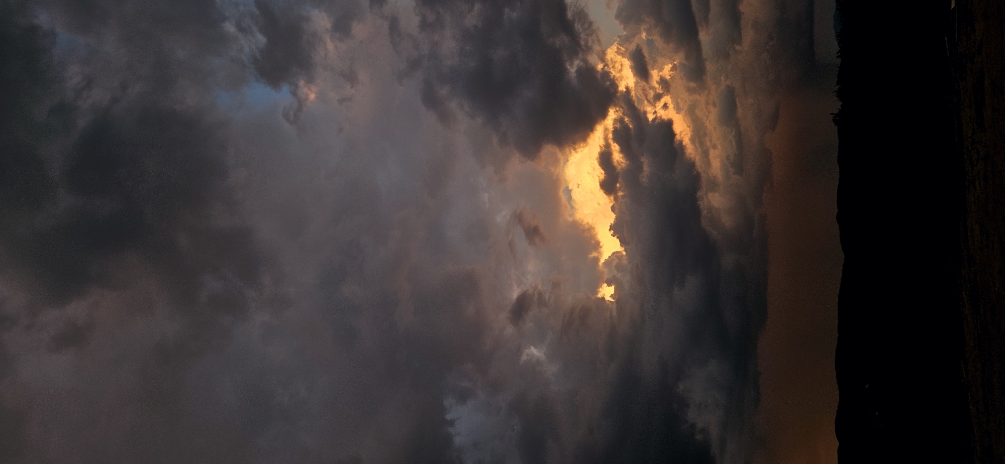 A dramatic, low-angle, vertical shot captures a stormy sky over the silhouette of hills in Momin Prohod, Bulgaria. The sky is filled with dark, heavy clouds, with a break in the center revealing a vibrant, glowing patch of orange and yellow light, suggesting sunset or sunrise. The light illuminates the edges of the surrounding clouds, creating a stark contrast. The foreground is a dark, silhouetted landscape of undulating hills, possibly with some sparse vegetation or distant structures, all rendered in deep shadow against the bright sky. The overall mood is one of awe and impending weather change.