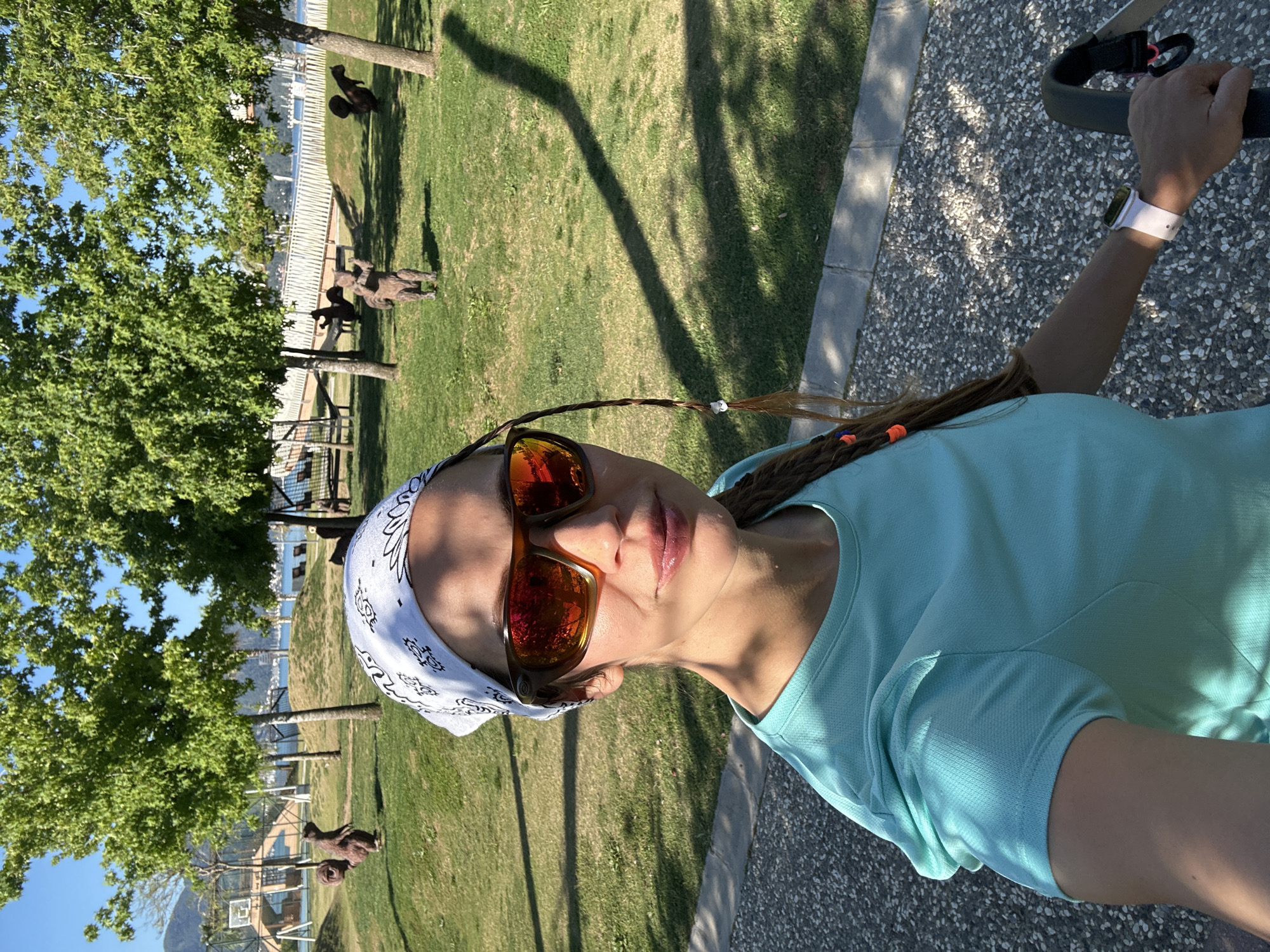 A woman is taking a selfie outdoors in Fethiye, Turkey. She has brown hair with some sections braided and secured with elastic hair ties. She is wearing a blue t-shirt, sunglasses with red lenses, and a white bandana. She is also wearing an Apple Watch. The background features a grassy area with trees. There are multiple animal statues, likely of goats, placed on the grass. The sea and a mountain are visible in the distance. A sidewalk is on the right side of the frame.