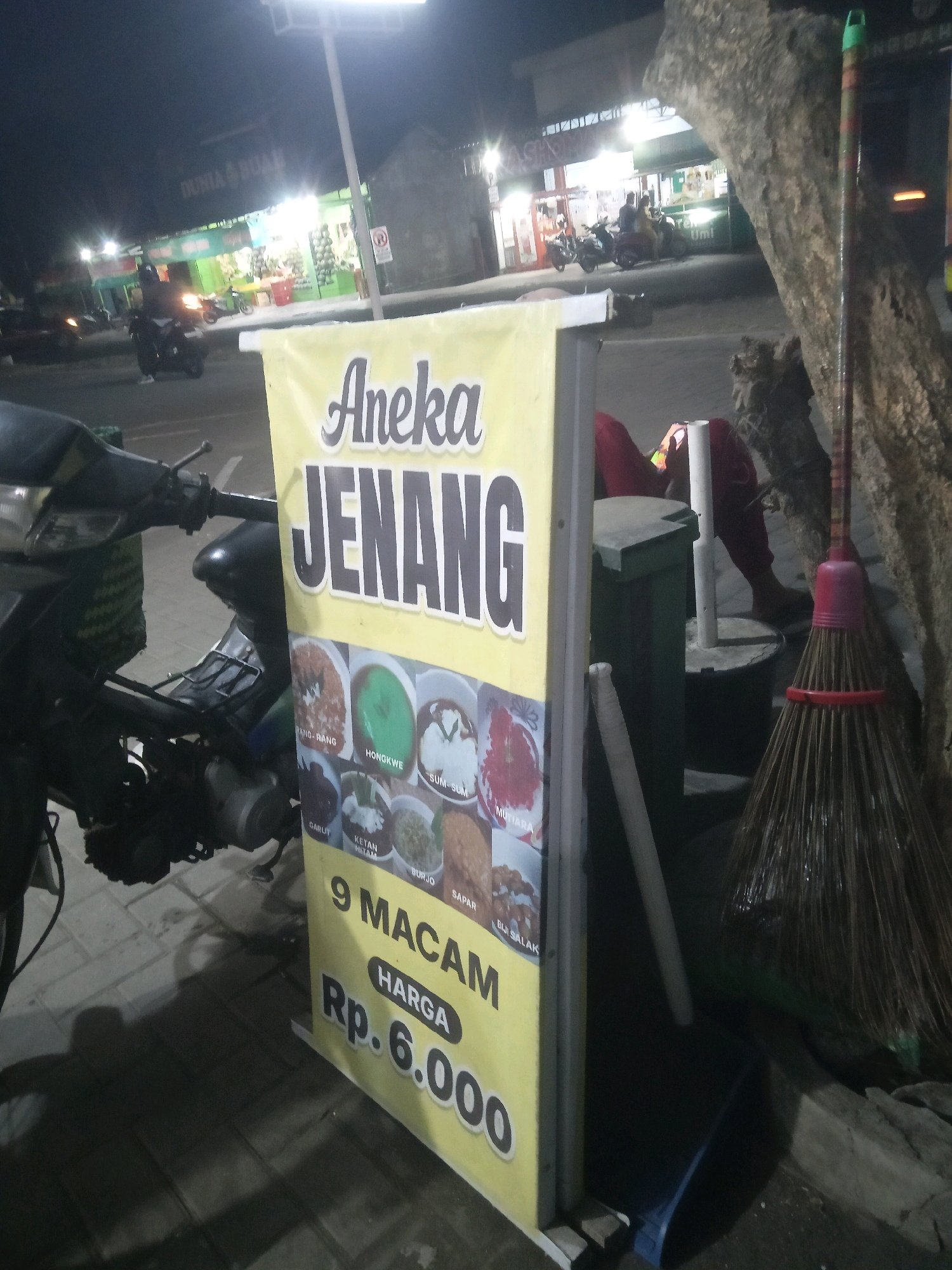 An outdoor scene at night, likely in Trihanggo, Indonesia, features a yellow banner advertising "Aneka Jenang" (various desserts) priced at Rp. 6.000 for 9 types. The banner is partially obscuring a parked motorcycle on the left. Behind the banner, a person in red trousers is seated near a green trash bin. To the right of the banner, a broom with a red handle leans against a tree trunk. In the background, several shops are illuminated, and motorcycles are visible on the street, suggesting a bustling commercial area. The lighting is dim, characteristic of nighttime.