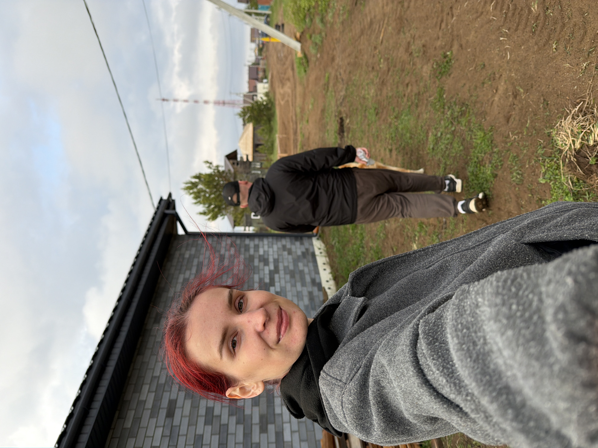 A sideways-oriented selfie captures a woman with red hair, wearing a gray fleece coat over a black hooded sweatshirt. Behind her, a person in a black jacket, brown pants, and a black cap walks a tan dog on a leash. The background reveals a dirt lot with sparse grass. In the distance, there is a cluster of houses, a cell tower, and an electrical pole. The side of a gray brick building is visible to the left. The location is the city of Chaikovsky, Russia.