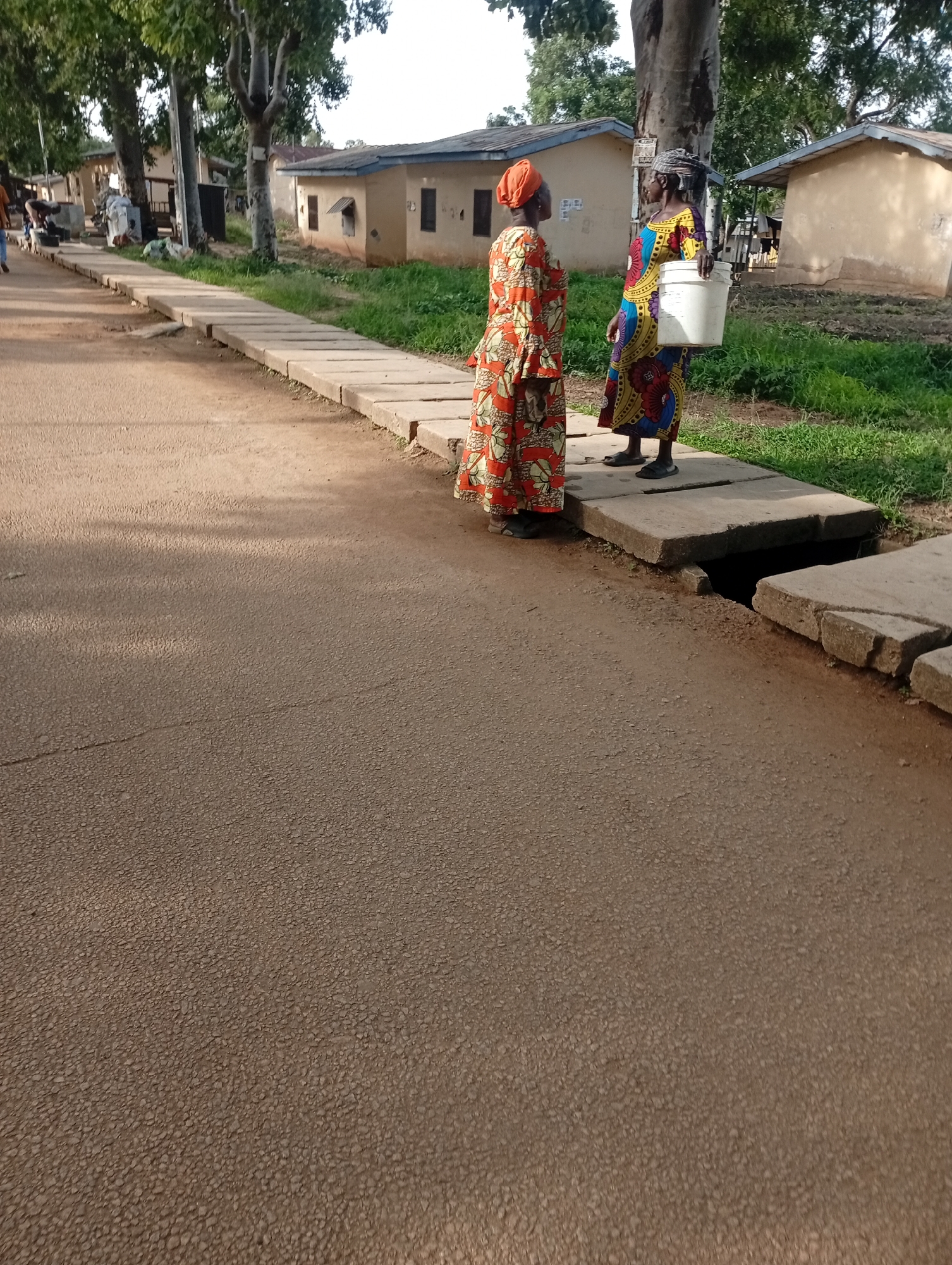 Two women stand on a concrete drainage cover next to a paved road in Rafin Par, Nigeria. The woman on the left wears a brightly colored orange and red patterned dress and an orange headscarf. She is facing away from the camera, looking to her left. The woman on the right wears a colorful, patterned dress in shades of blue, yellow, red, and black, and a dark headscarf. She is holding a large white bucket in her right hand and is facing the woman in the orange dress. Behind them are two low-lying buildings with light-colored walls and dark roofs. The environment is rural, with trees and overgrown grass visible. The road appears to be asphalt and is in the foreground, with a concrete sidewalk or drainage system running alongside it. The overall lighting suggests it is daytime, possibly late afternoon due to the shadows cast by the trees.