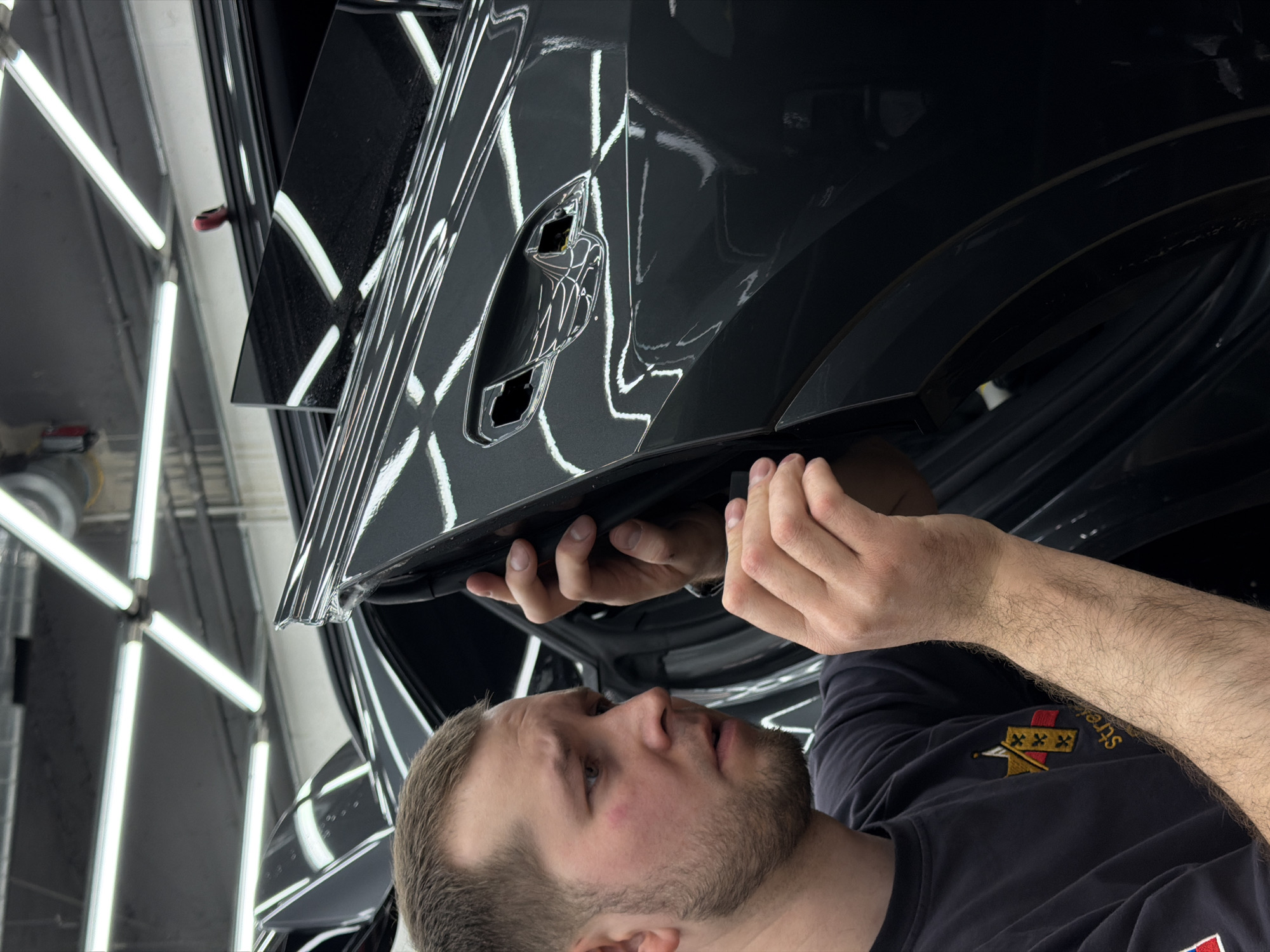 A Caucasian man is depicted in an indoor environment, focused on affixing an object to the side of a vehicle. The man has short, light brown hair and a beard. He is wearing a dark-colored t-shirt. He appears to be concentrating on the task at hand.
The vehicle is dark and highly reflective, presumably due to a recent detailing or coating. The area surrounding the vehicle and the man is brightly lit by a series of overhead fluorescent lights.
The location is unknown. There is no identifying information in this image to suggest the location is Krasnoyarsk, Russia.