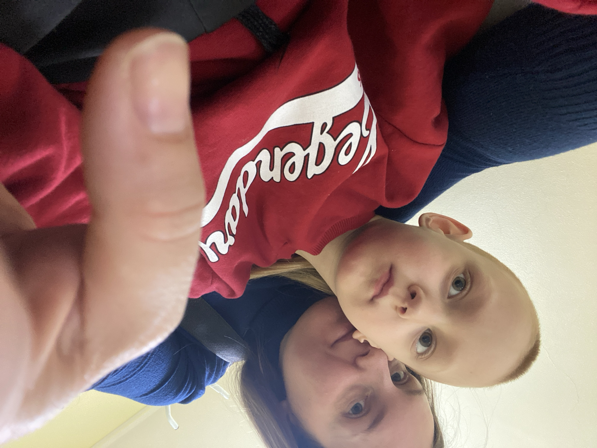 A selfie taken from a low angle shows a woman and a child in what appears to be an indoor setting. The child is wearing a red sweater with the word "Legends" in white script. The woman is wearing a blue top. A thumb is visible in the foreground, slightly out of focus. The background is a pale yellow, likely a wall. The location and city cannot be determined from the image.