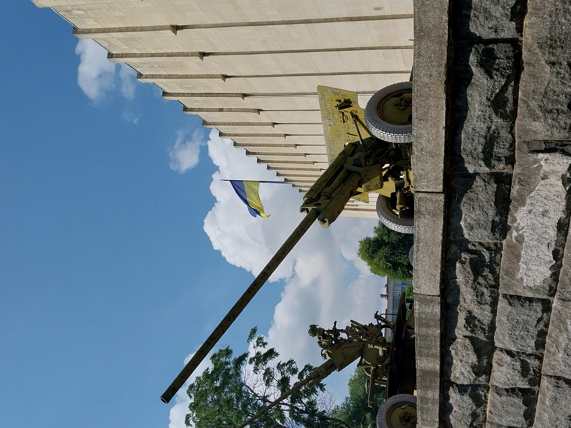 The image depicts two green artillery pieces positioned on a stone platform in front of a light-colored, modern building with vertical architectural elements. A Ukrainian flag, featuring blue and yellow colors, is visible flying in the background between the two cannons. Trees are present to the left and behind the artillery pieces. The sky is blue with scattered white clouds. The scene is set outdoors, with the building possibly being a museum or memorial. This location is consistent with the monument in the Dnipro city, Ukraine, dedicated to the defenders of the city.