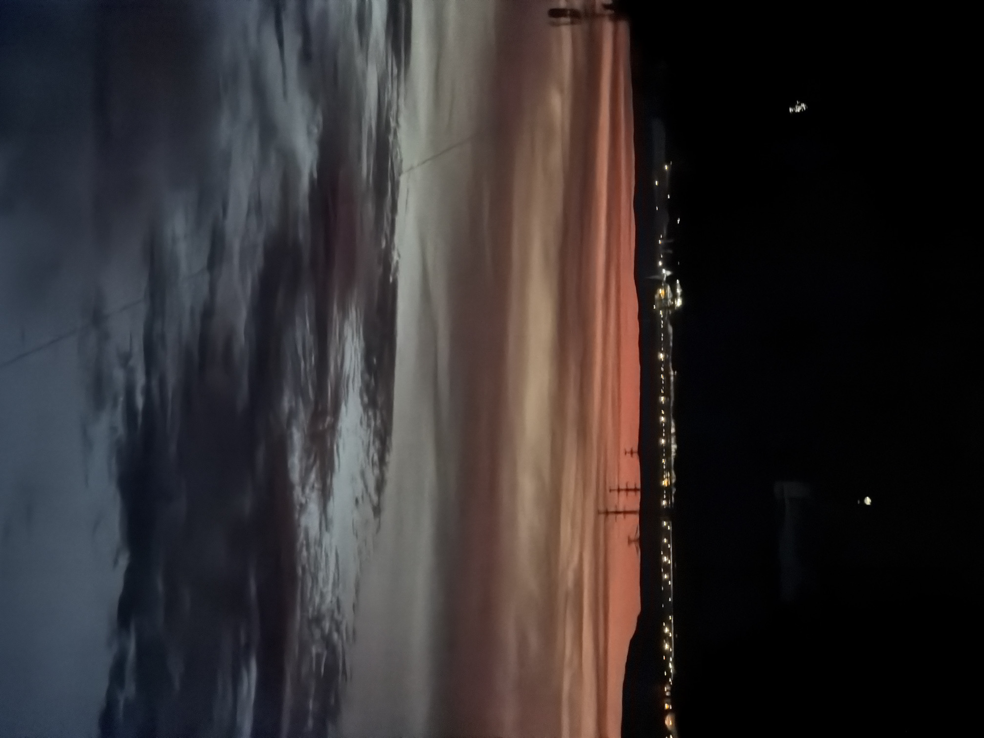 A wide shot captures a darkening sky over Chaikovsky, Russia. The upper portion of the sky is a deep, dusky blue, transitioning to bands of orange and pink as it nears the horizon. Wispy clouds streak across the sky, with some appearing darker and more defined. Silhouetted against the horizon are hills and a distant cityscape illuminated by numerous small lights. Three tall, lattice-like power transmission towers stand prominently in the mid-ground. The foreground is predominantly dark, obscuring details, but the faint outline of a building or structure is visible in the lower left. The overall impression is of dusk or twilight in an industrial or semi-urban area.