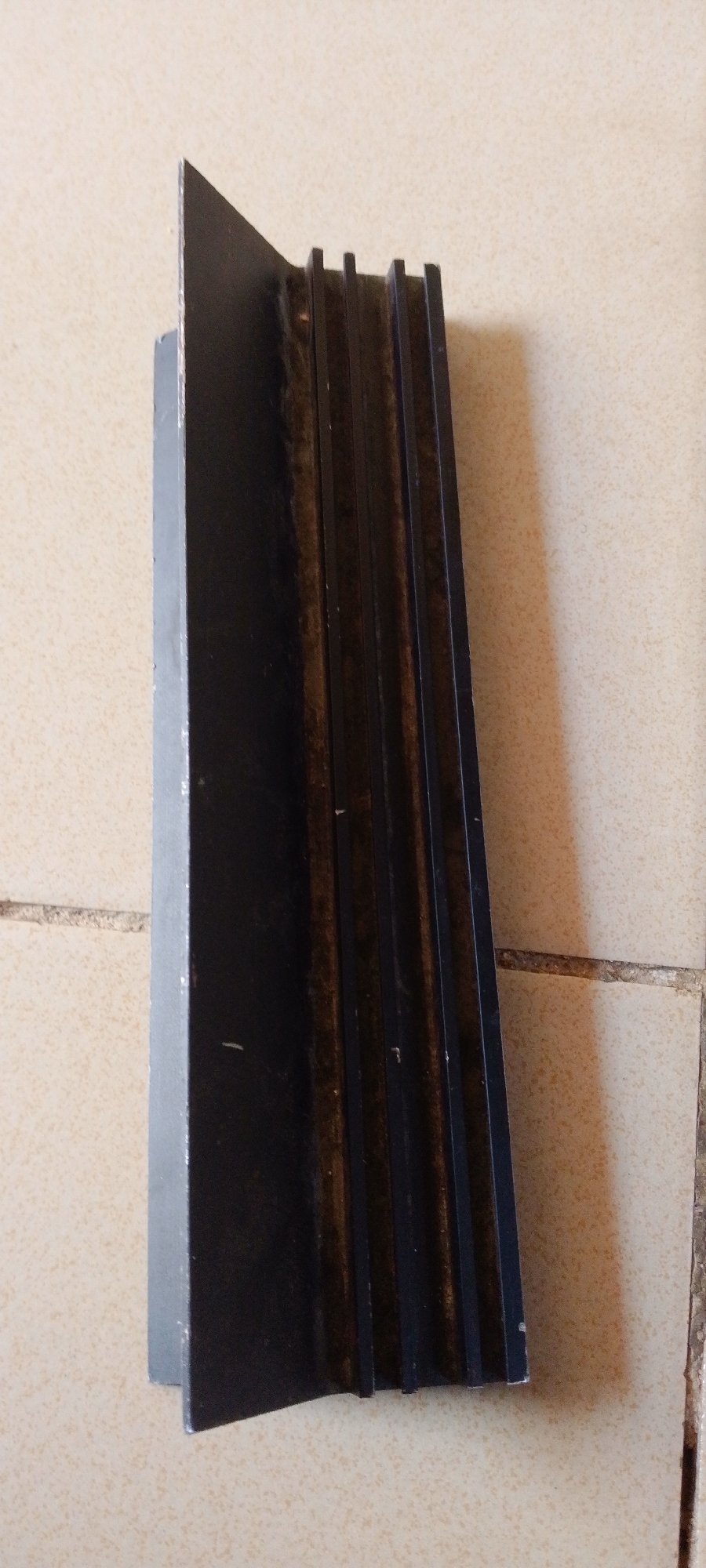 This is a close-up, eye-level photograph of a black, multi-grooved object placed on a textured, light-colored tiled surface. The object appears to be made of metal and has a sharp, angled edge on its left side. The right side of the object features several parallel grooves or fins, which suggest it might be a heat sink or a decorative element. The object is positioned diagonally across the frame, with its pointed end facing towards the top left. The tiled surface is light beige or cream-colored and has visible grout lines. The lighting suggests it is daytime, with soft shadows cast by the object onto the tiles. The overall scene is simple and focuses on the texture and form of the object and its surroundings. There is no visible text in the image. The location context provided is Jalingo, Nigeria.